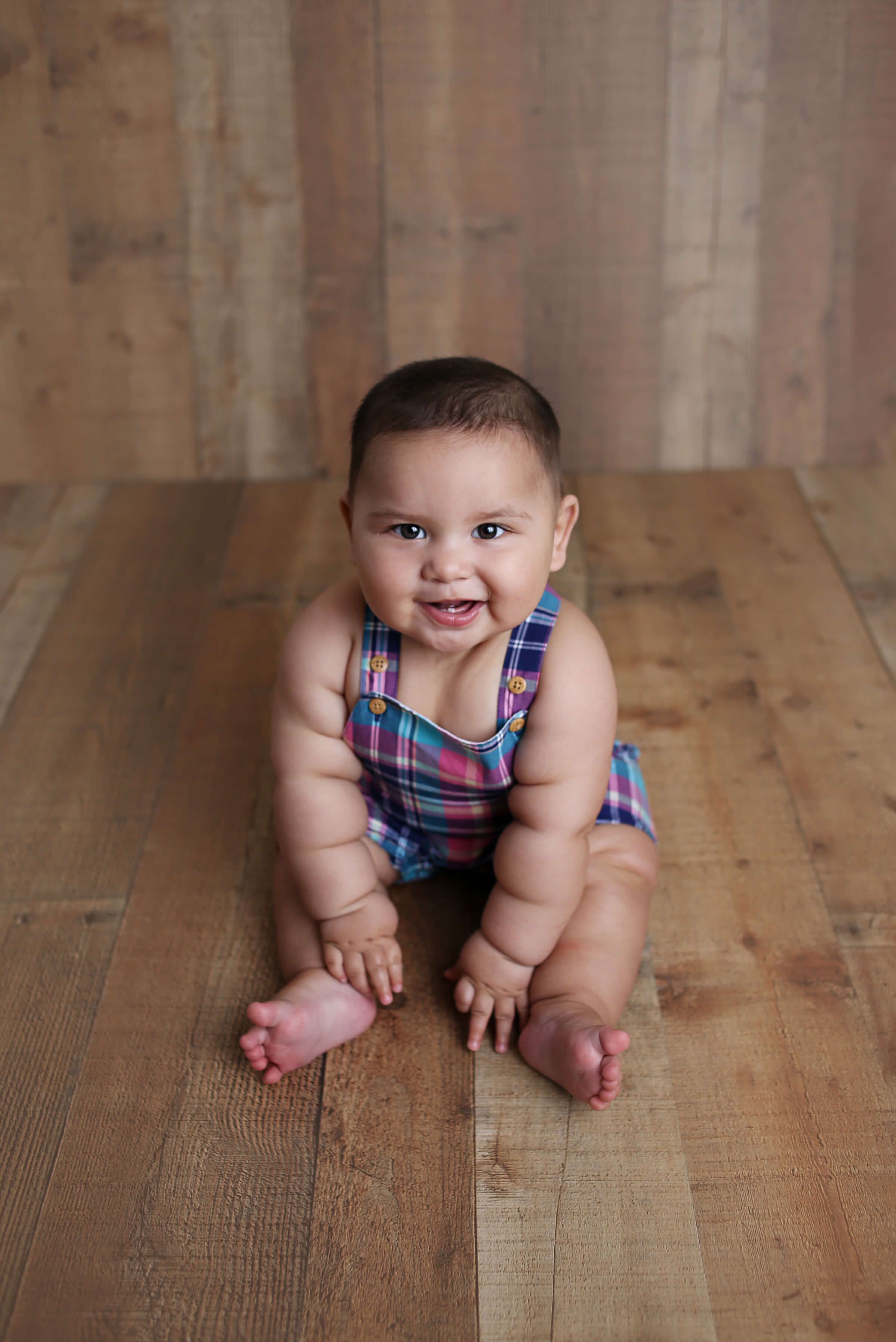  A photograph of a smiling baby boy, sitting in a bright plaid playsuit, with a simple wooden backdrop, celebrating a one-year-old milestone from an infant picture session 