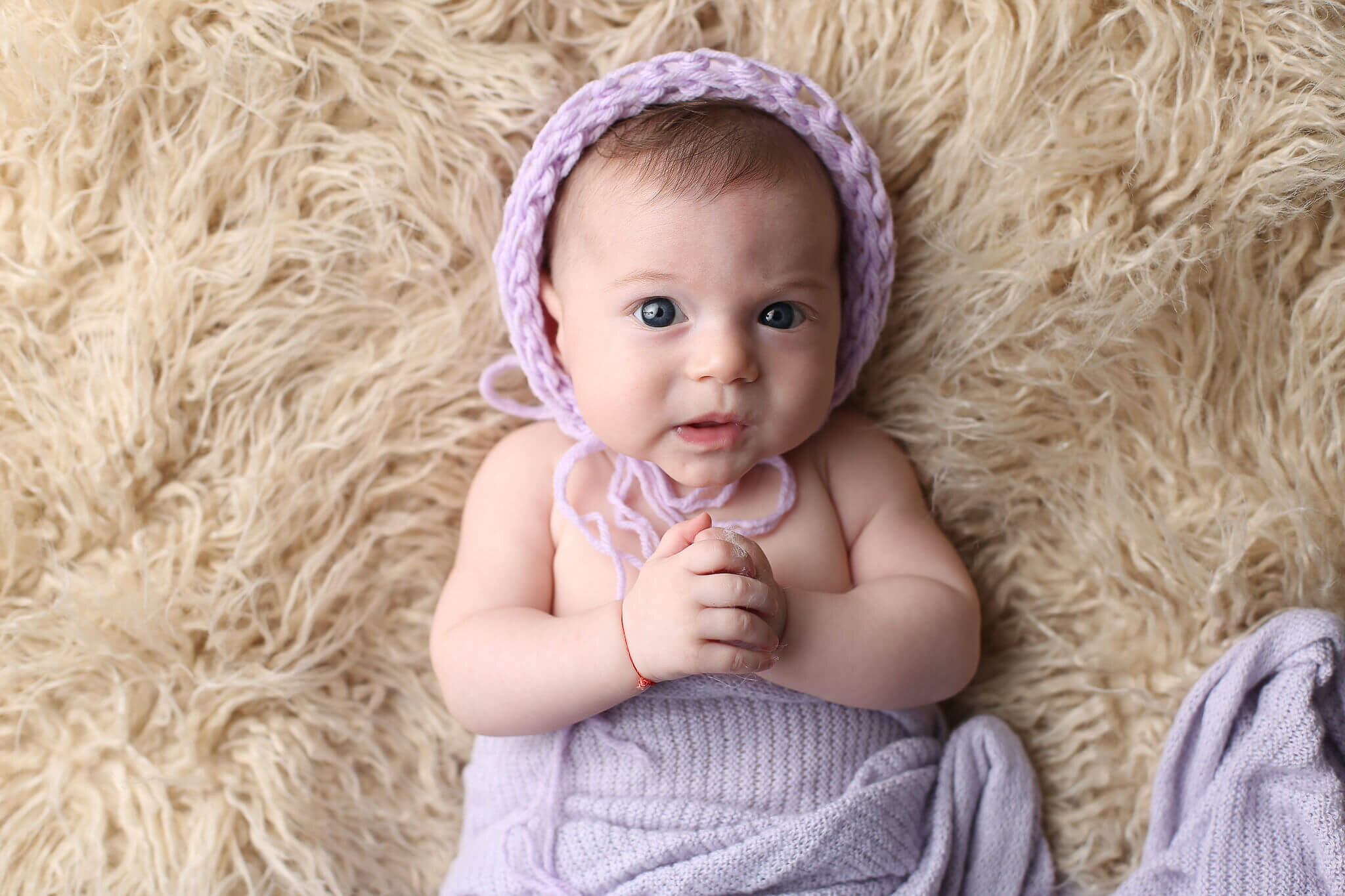  A picture of a tiny baby girl in her knitted cap and blanket, lying on a shaggy rug and holding her hands together as she looks up with her sweet face by Photography by L Rose, baby photography in San Diego CA 