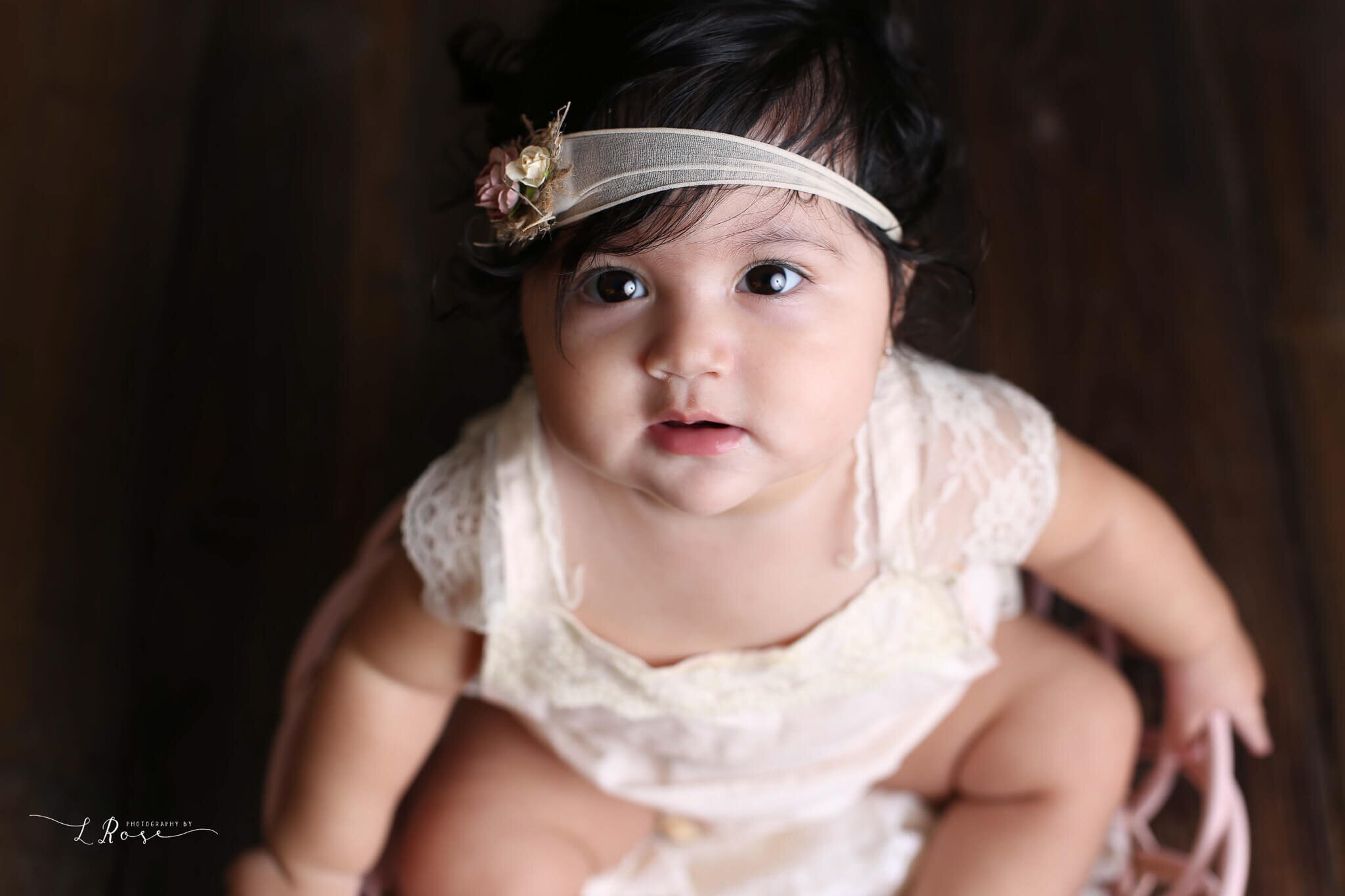  A picture from above of a sweet girl in a flowered headband and lacy dress, looking up with her dark eyes as she celebrates a milestone from a baby photo session 