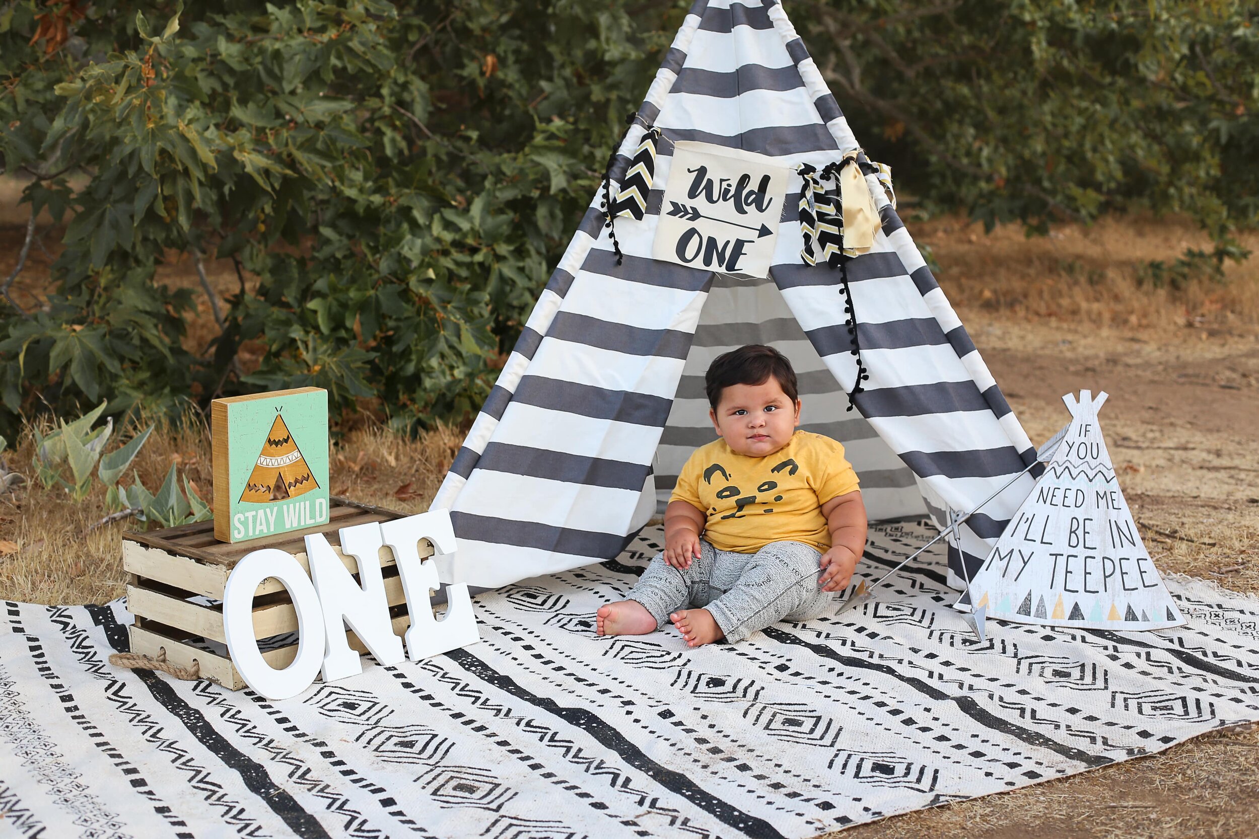 A picture of a cute boy in his play clothes sitting in front of a play tent, celebrating his one-year-old milestone with big letters spelling out “one” by Photography by L Rose, baby photography in San Diego CA 