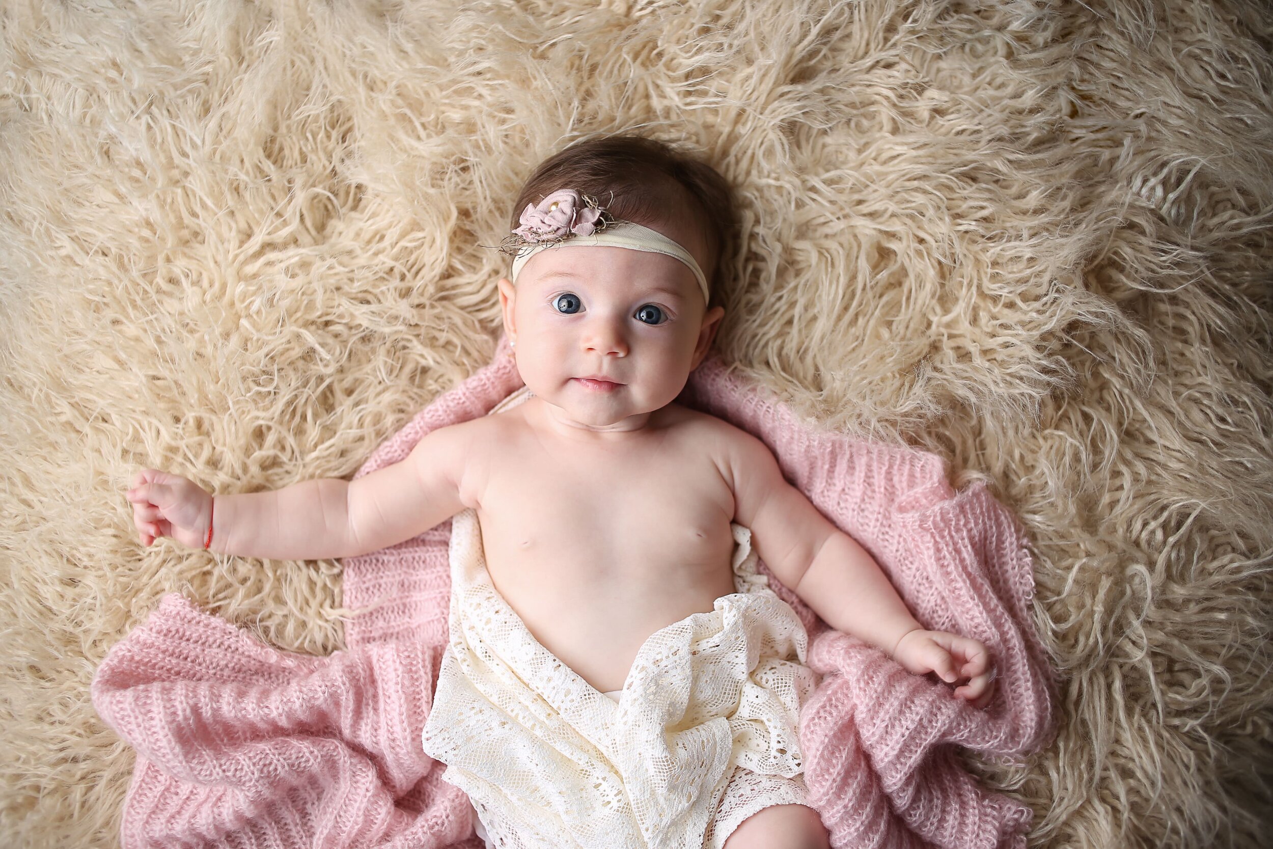  A photograph from above of a sweet baby girl in a flowered headband, looking up with a happy face as she is wrapped in a lacy cover and pink blanket, reaching a new milestone by Photography by L Rose, a baby photographer in San Diego California 