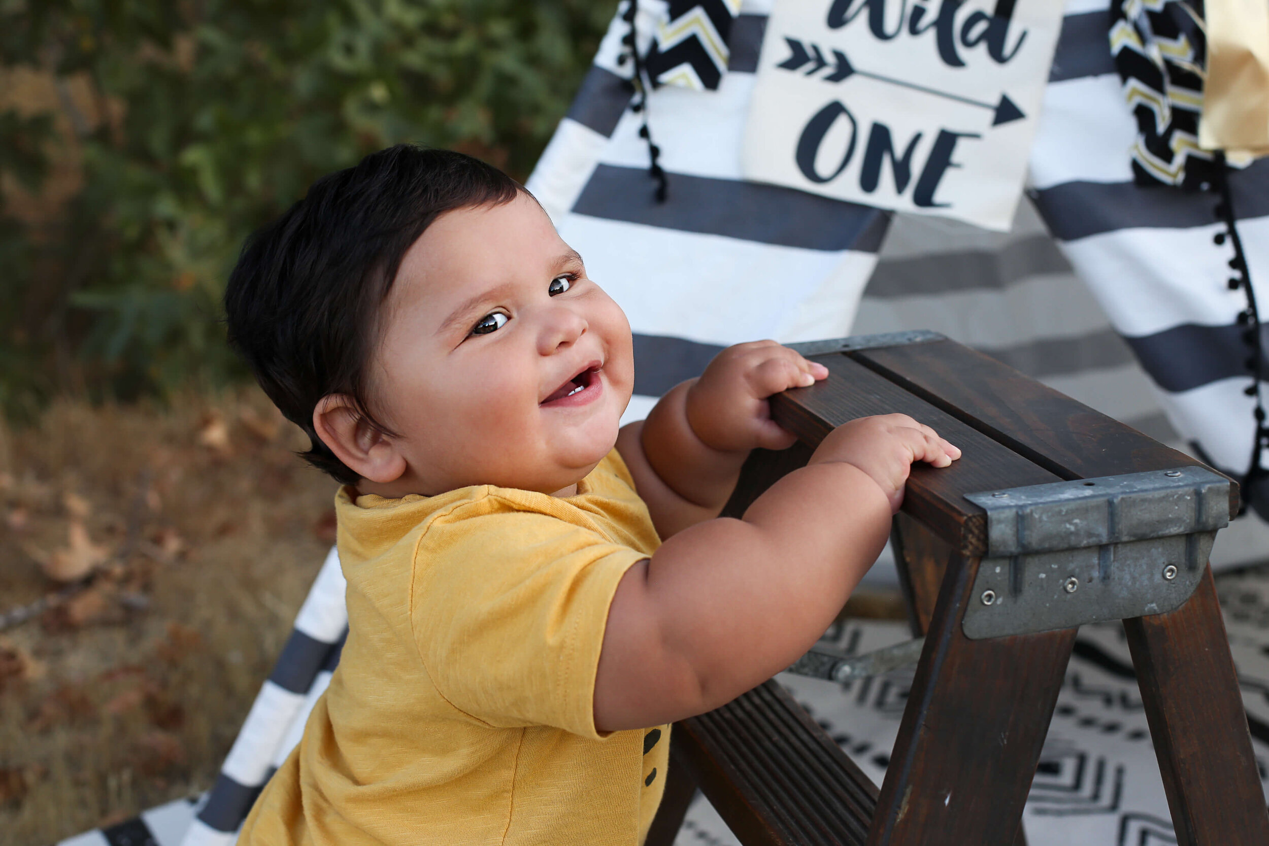  An image of a closeup view of a smiling one-year-old boy holding himself up on a stepstool, reaching a new milestone from a baby milestone photo session 