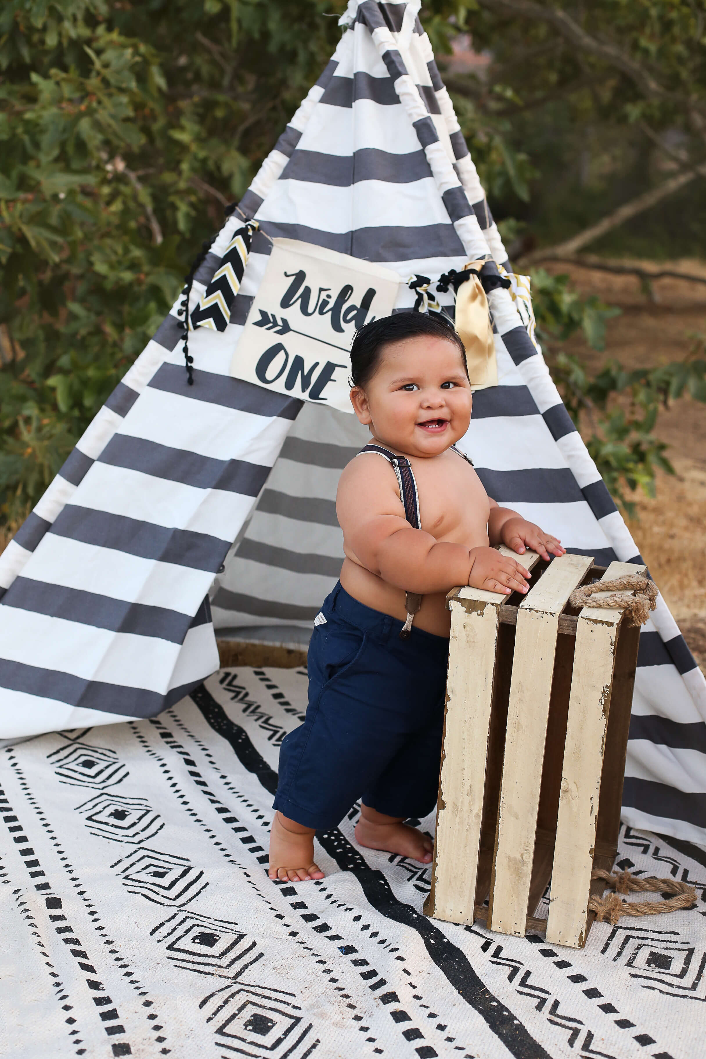  A photograph of a happy one-year-old boy in pants and suspenders, standing and holding onto a vintage crate in front of a cute little Indian teepee as he reaches a milestone from an infant picture session 