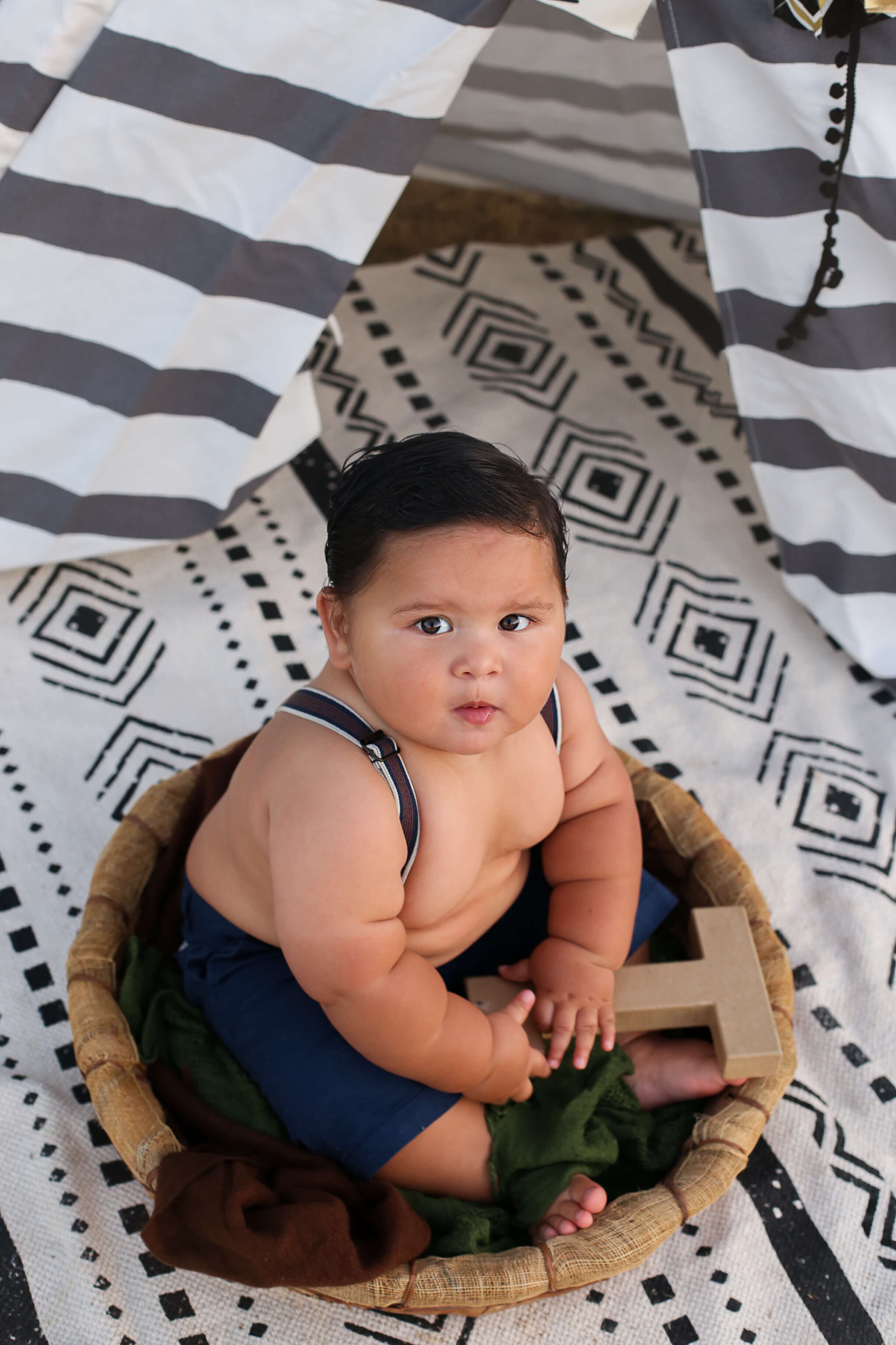  An image from above of a boy sitting in a rustic basket, holding a large letter that represents his name, as he celebrates his one-year-old milestone by Photography by L Rose, baby photography in San Diego 