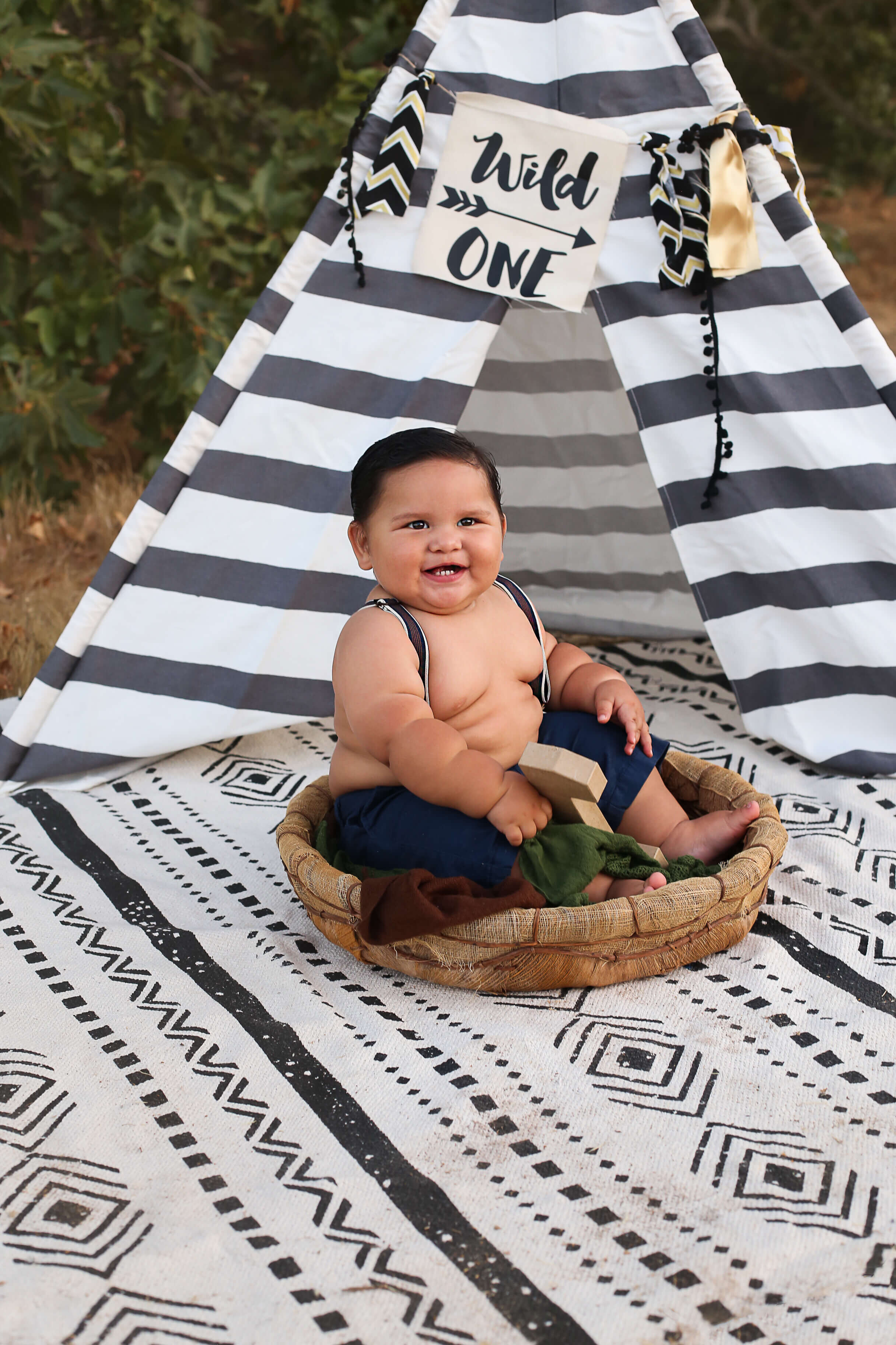  A picture of a one-year-old boy smiling and sitting in a cute basket in his pants and suspenders, in front of a little Indian teepee, marking a milestone from a baby photo session 