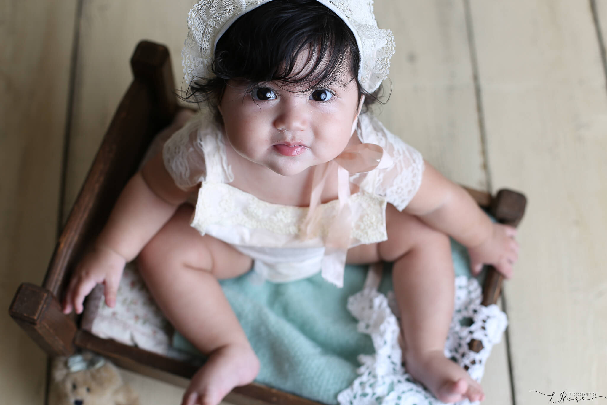  A photograph from above of a cute baby girl wearing a lacy bonnet and playsuit, sitting in a small cradle as she looks up at the camera, marking a milestone by Photography by L Rose, a baby photographer in San Diego California 