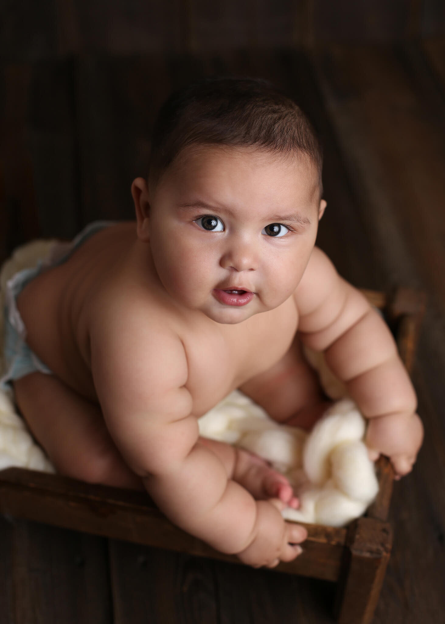  A picture from above of a cute baby boy sitting on a soft blanket, leaning over and looking up at the camera, reaching a new milestone from a baby photo session 