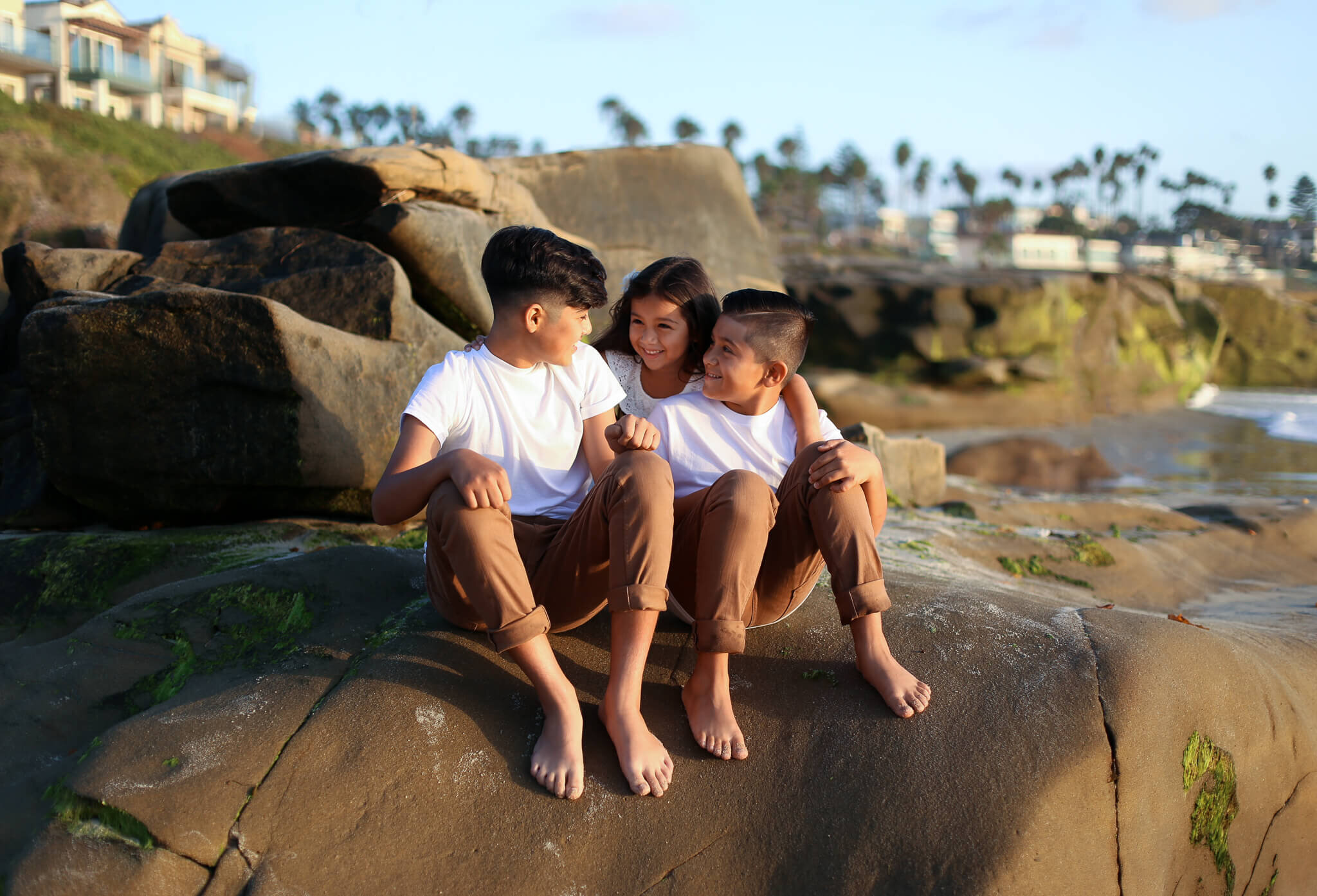  An image of three young siblings sitting in their bare feet close together on some rocks, smiling in the sunshine during family time together outdoors by Photography by L Rose 