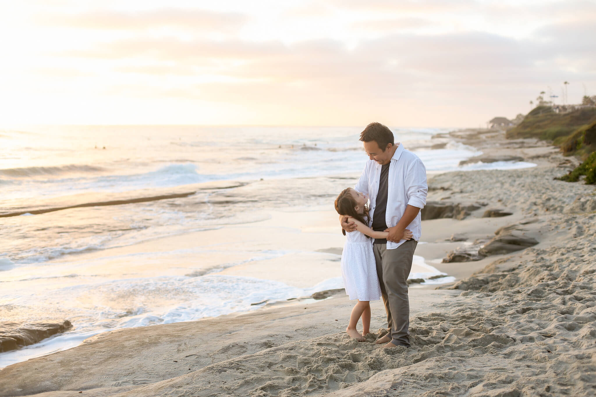  A photograph of a father gazing down at his young daughter, holding her close, as they stand on the beach at the water’s edge, spending family time together from a family photo gallery 