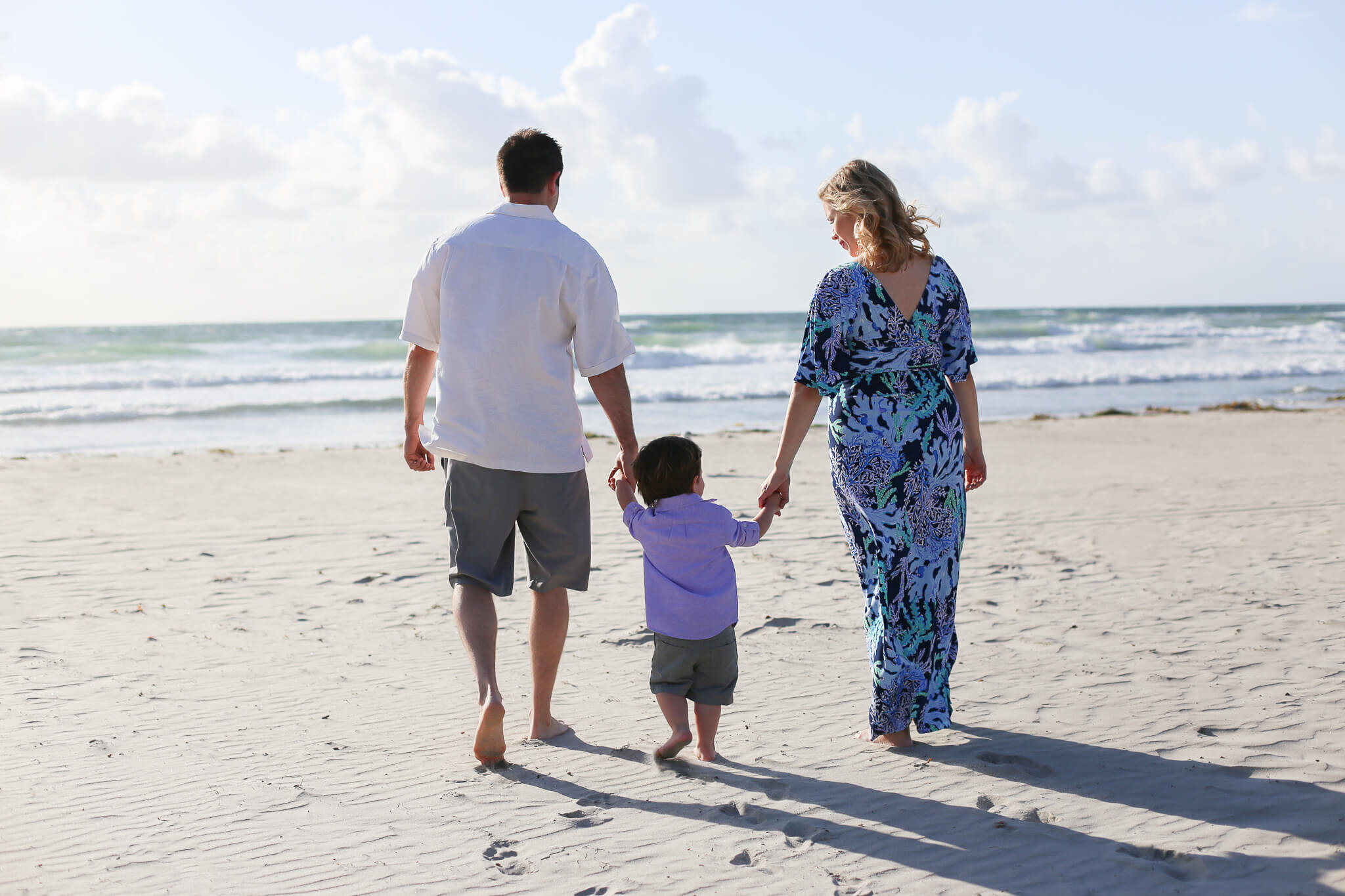  An image of a mother and father from behind, holding hands with their young son between them as they stroll as a family along the beach by the ocean on a sunny day 