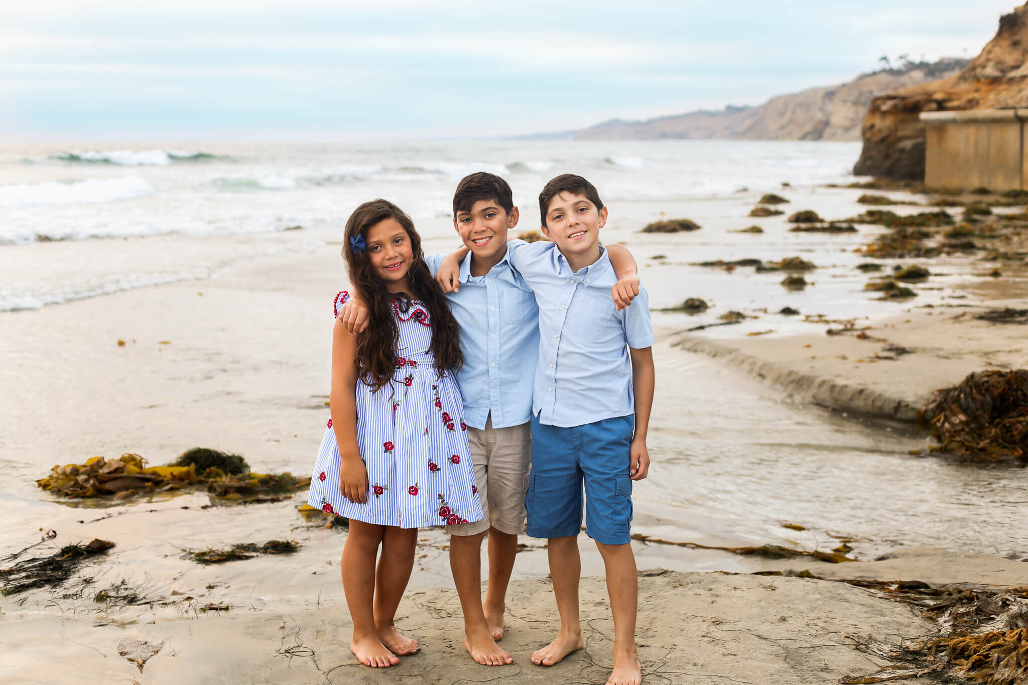  An image of three young siblings standing with their arms around each other, smiling as they spend the day at the seashore with their family in a family photo session 