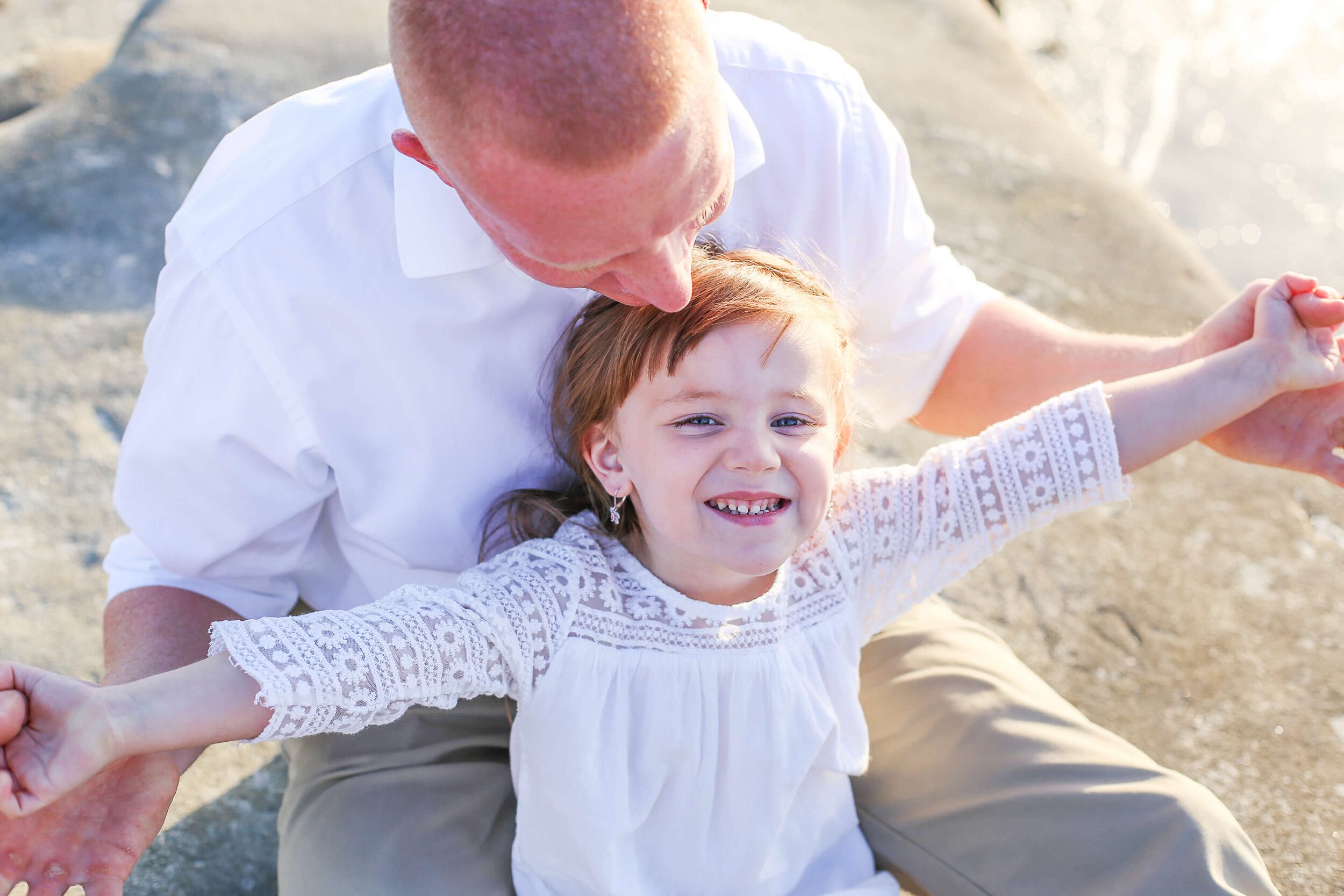  An image of a dad sitting with his young daughter in the sand as he playfully holds her arms out, relaxing together as a family 