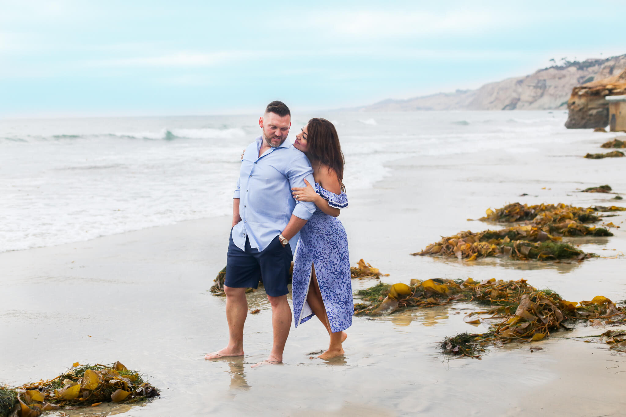  A picture of a wife romantically placing her chin on her husband’s shoulder from behind as they stand together at the ocean’s edge, spending family time together outdoors 