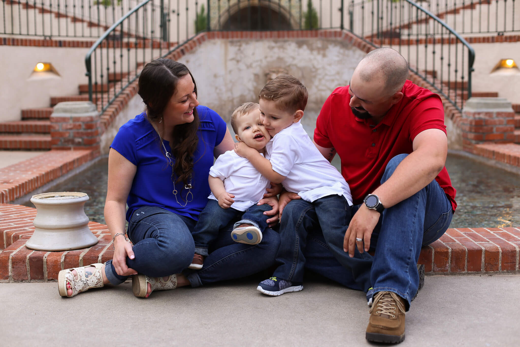  An image of a family sitting together in front of a vintage building as the two young brothers hug each other and the mom and dad look on lovingly in a family photo session 