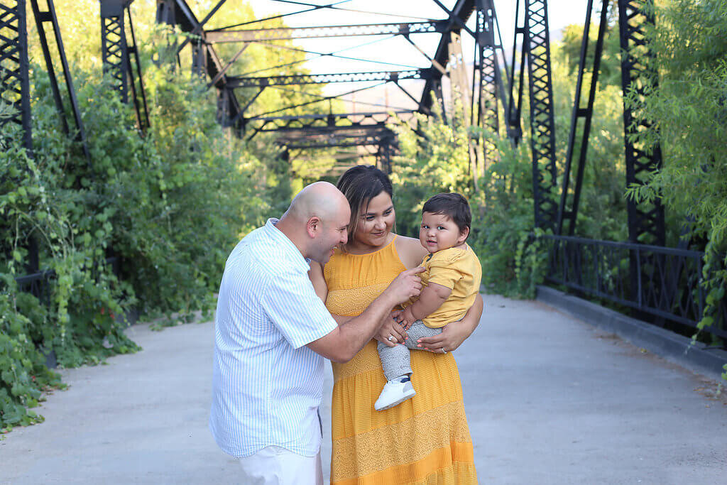  A picture of a family standing close together on an old bridge as the mother holds their son in her arms while dad tickles him from a family pictures photo session 