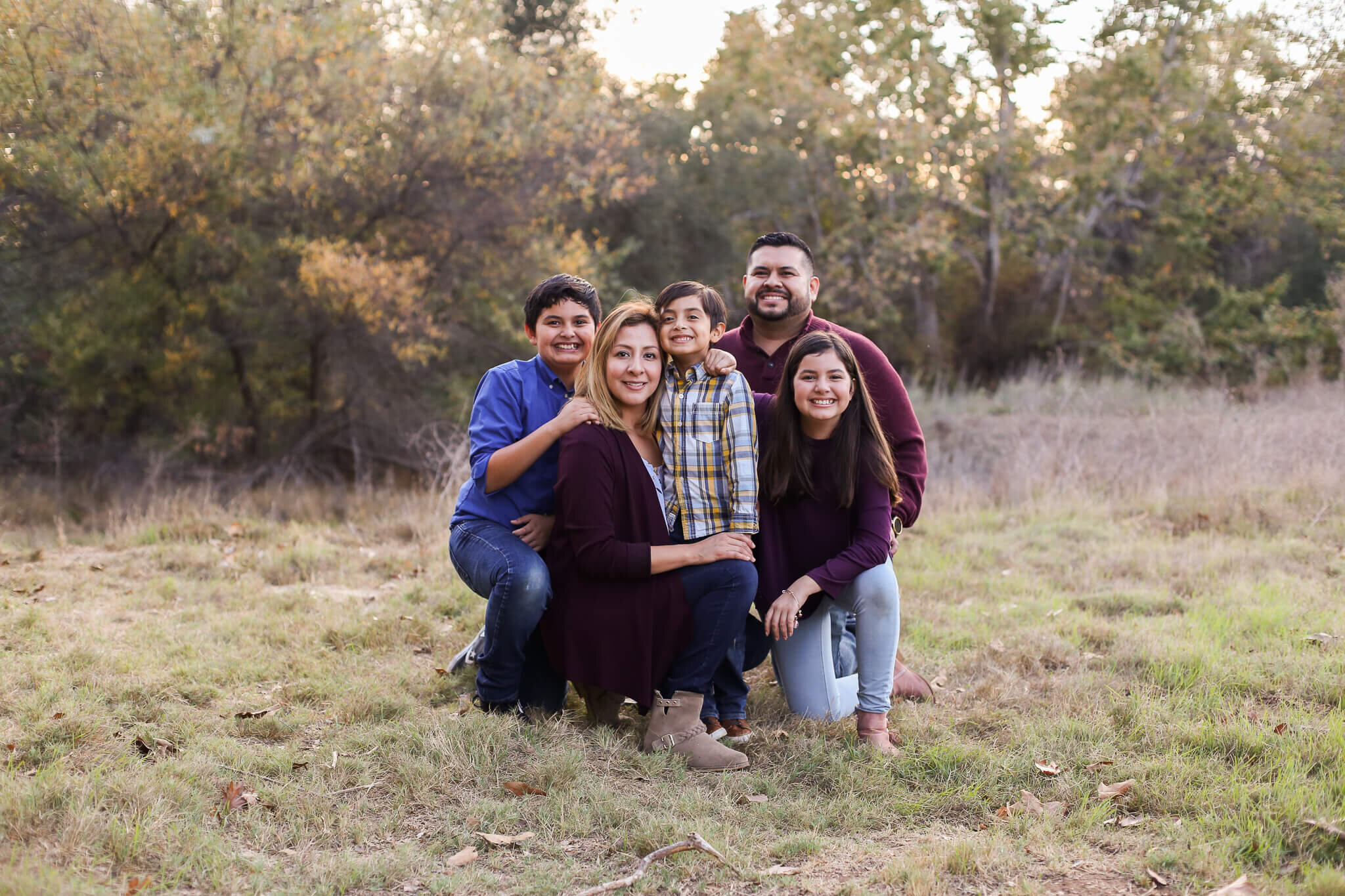  A photograph of a family kneeling down close together in the grass as they smile and hold each other close, spending time together outdoors by Photography by L Rose 