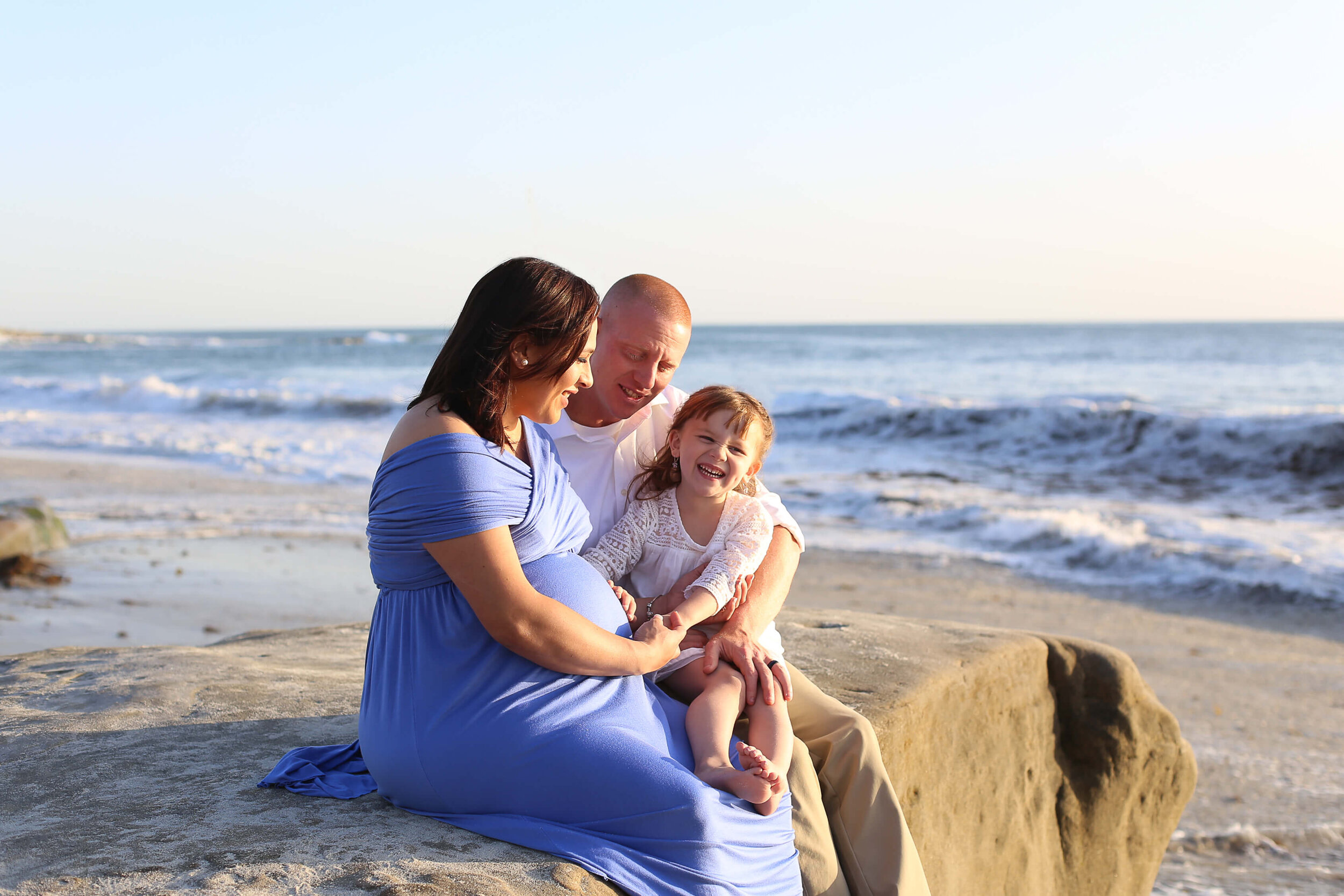  A photograph of a family sitting on some rocks together by the ocean, as the young daughter smiles and touches her mother’s baby bump by Photography by L Rose - San Diego California family photographer 