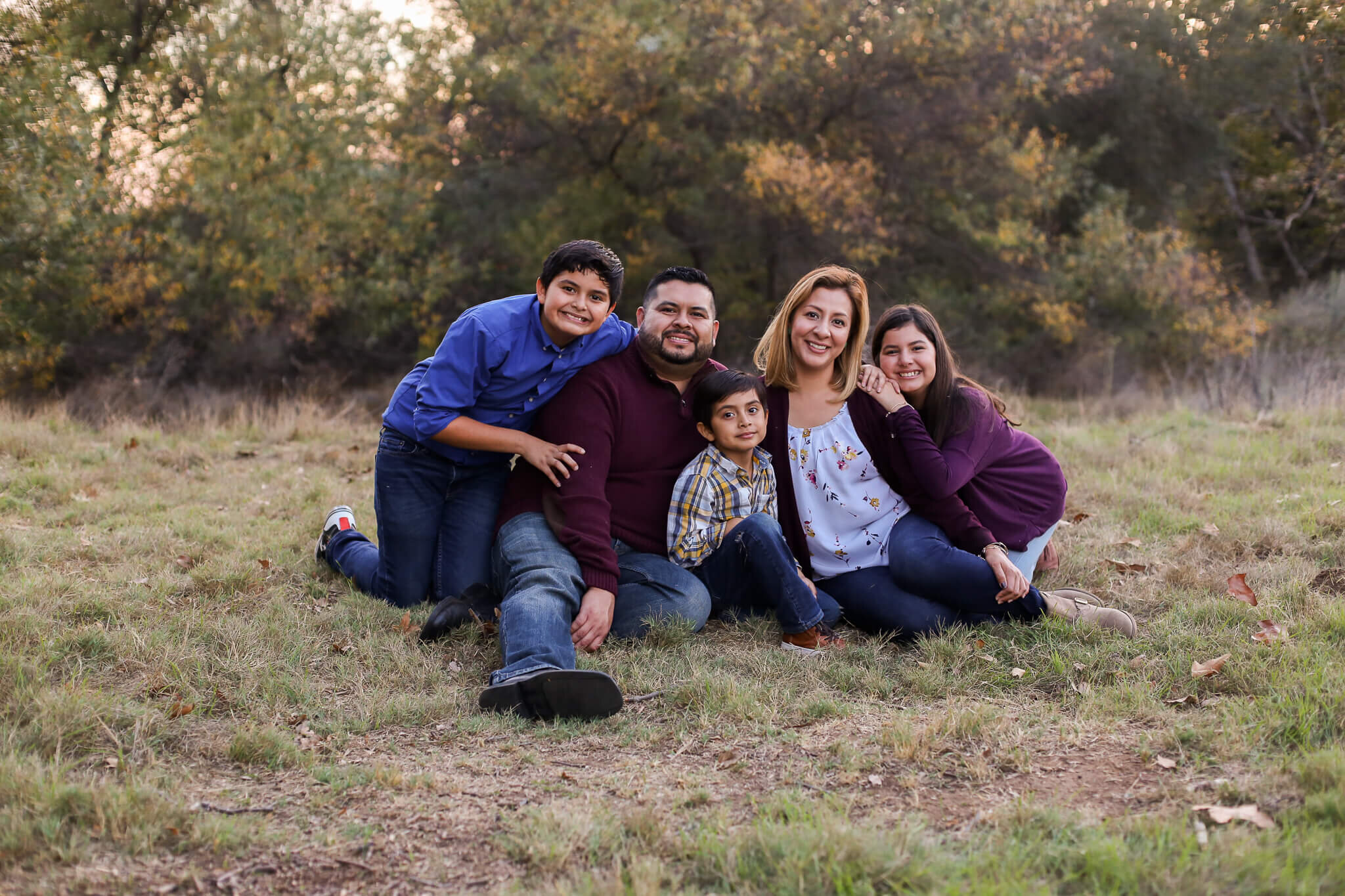  An image of a family sitting on the grass together as they smile warmly, holding each other close, enjoying their time together in a family photo session 