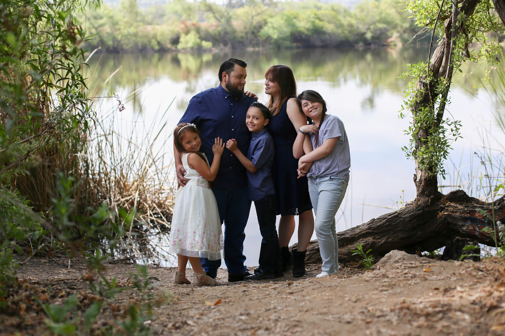  A photograph of a family standing close and hugging each other as the mother and father gaze lovingly at each other while the children smile happily, standing by a peaceful river from a family photo gallery 