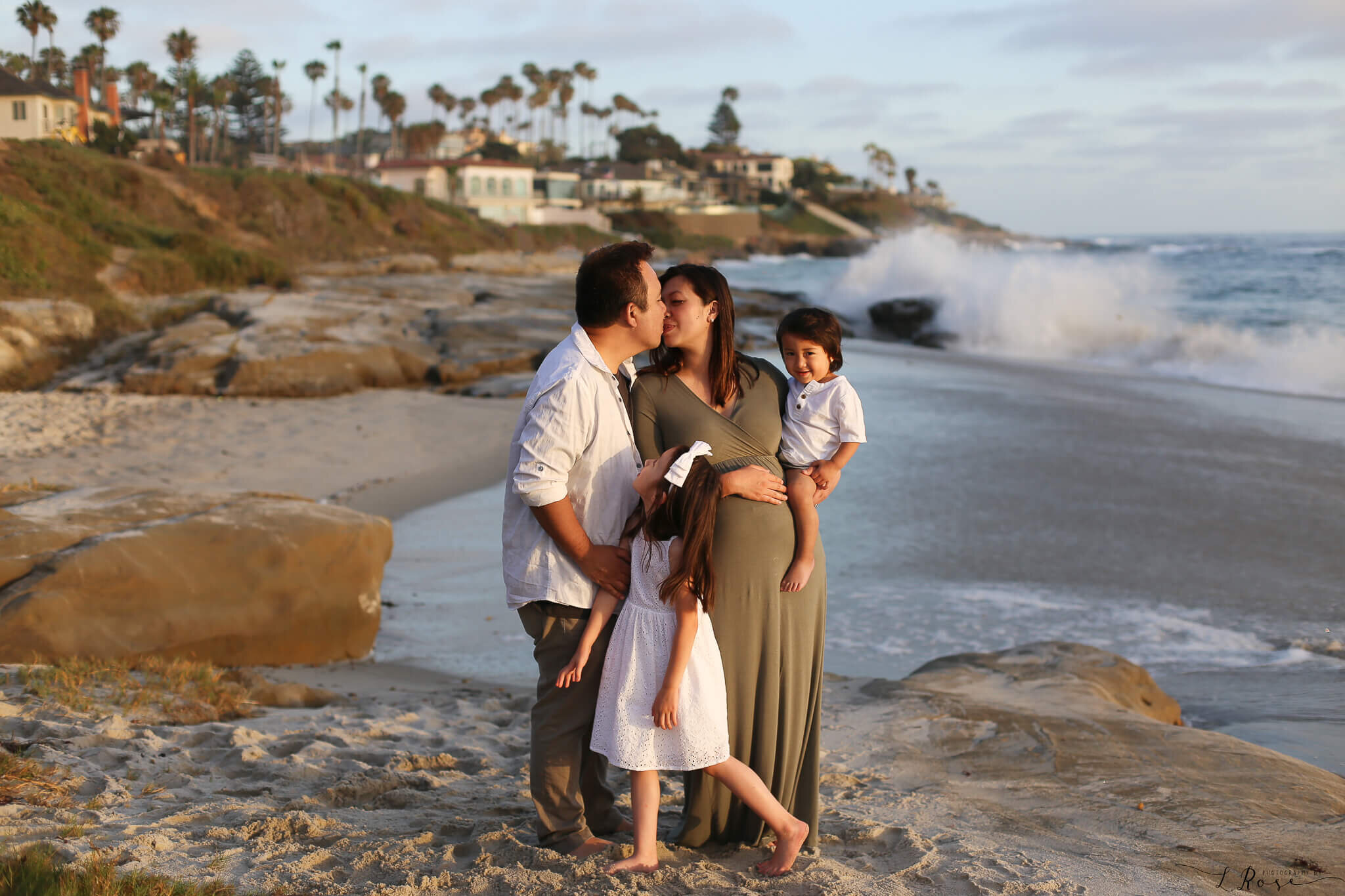  A photograph of a loving family as the expectant mom and dad romantically kiss as they hold onto their two children, standing at the edge of the ocean with a picturesque shoreline behind them from a family photo gallery 