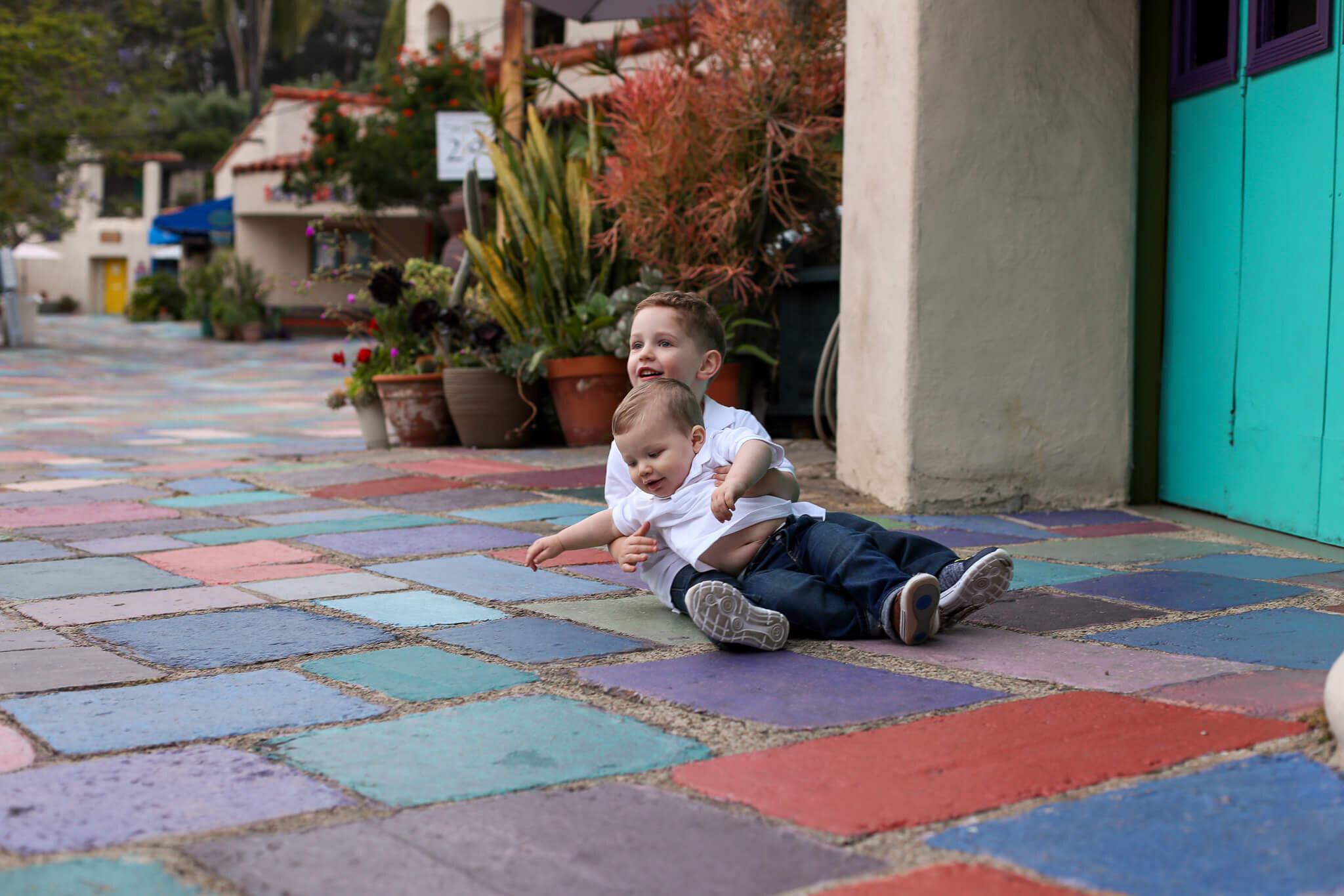  An image of two young siblings having fun wrestling together on a colorful outdoor tiled patio, spending time on a family outing by Photography by L Rose 