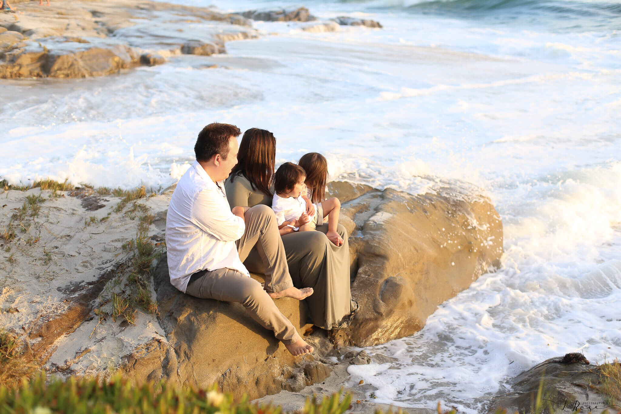  A picture of a family sitting close together on a rock near the ocean’s crashing waves, enjoying time together in the sunny weather from a family pictures photo session 