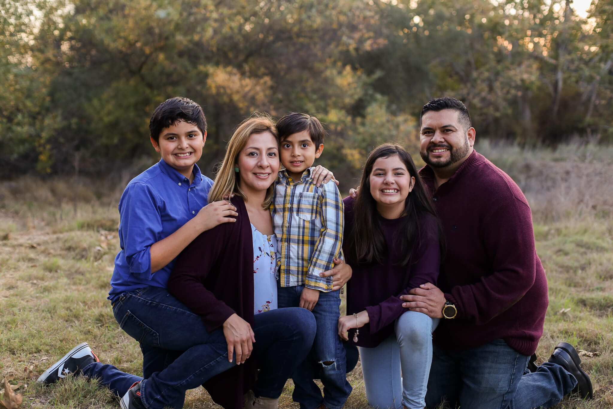  A picture of a closeup view of a family kneeling down, embracing each other as they lovingly enjoy their time in a park surrounded by trees 