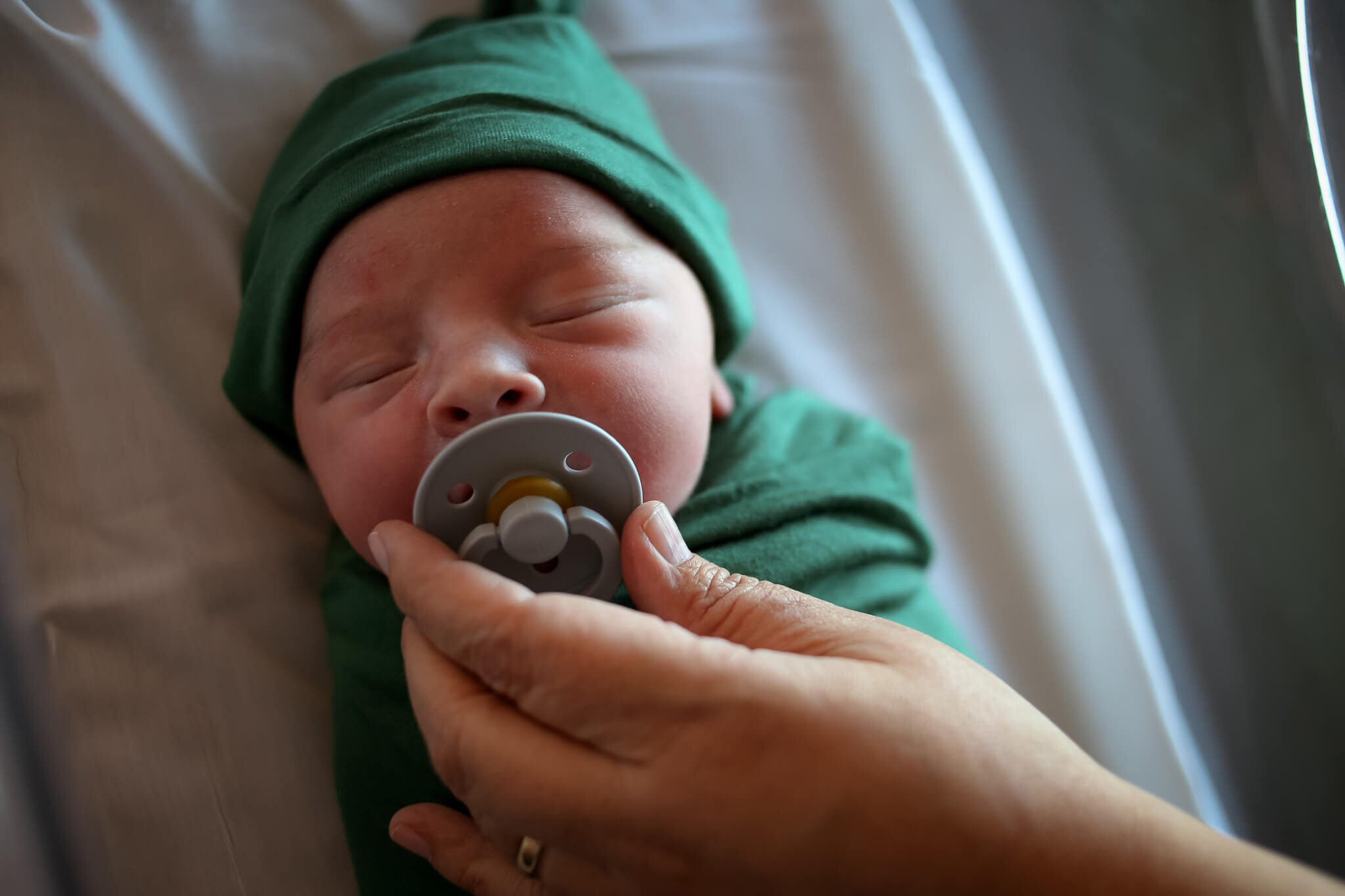  A picture of a closeup view of a newborn in a cap and tightly bound in a blanket with mom’s hand holding a pacifier in place as the tiny infant sleeps by Photography by L Rose - San Diego CA newborn photography 