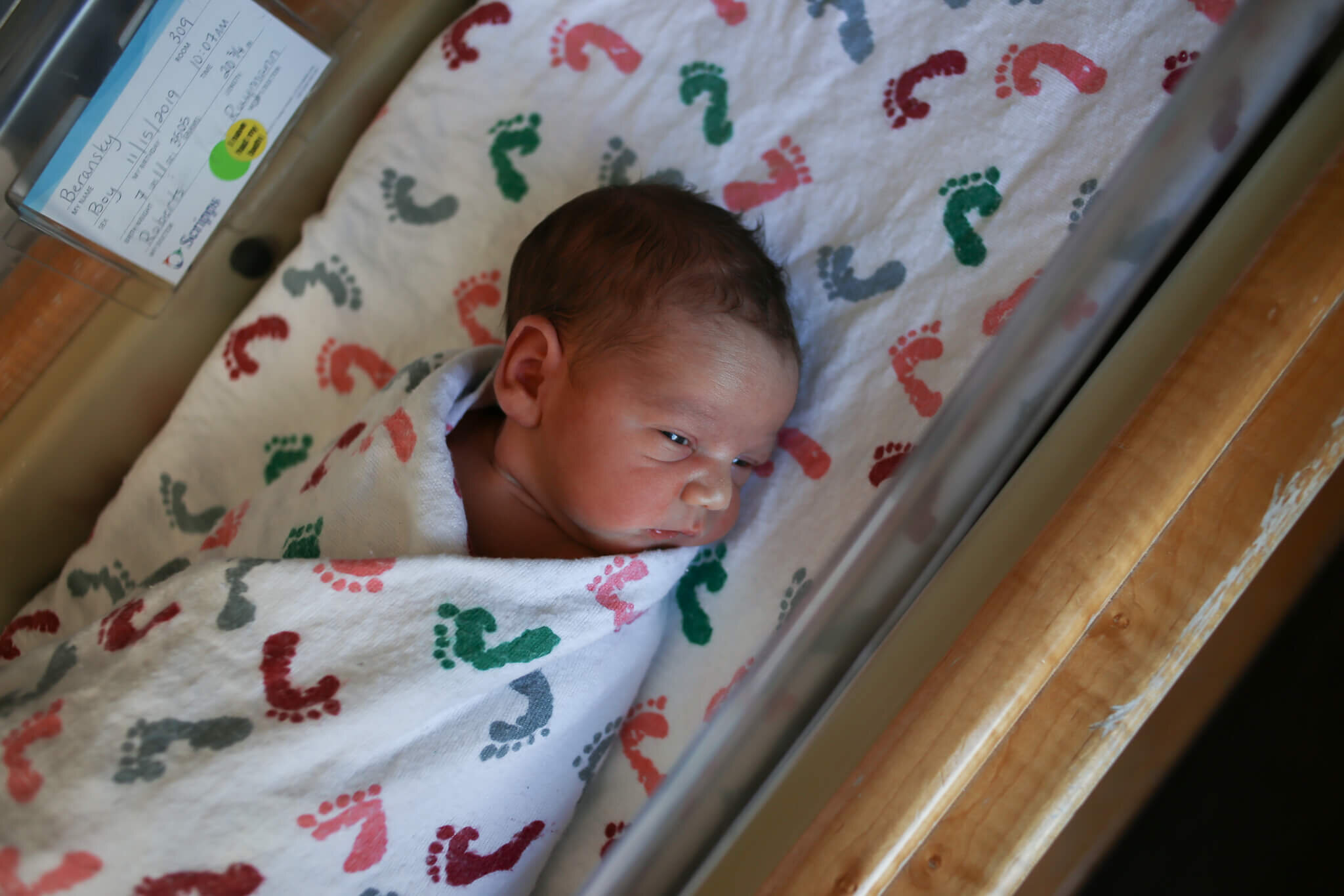  A photograph from above of a newborn baby with his eyes half-open, wrapped in a blanket and lying in a hospital bassinet from a newborn picture session 
