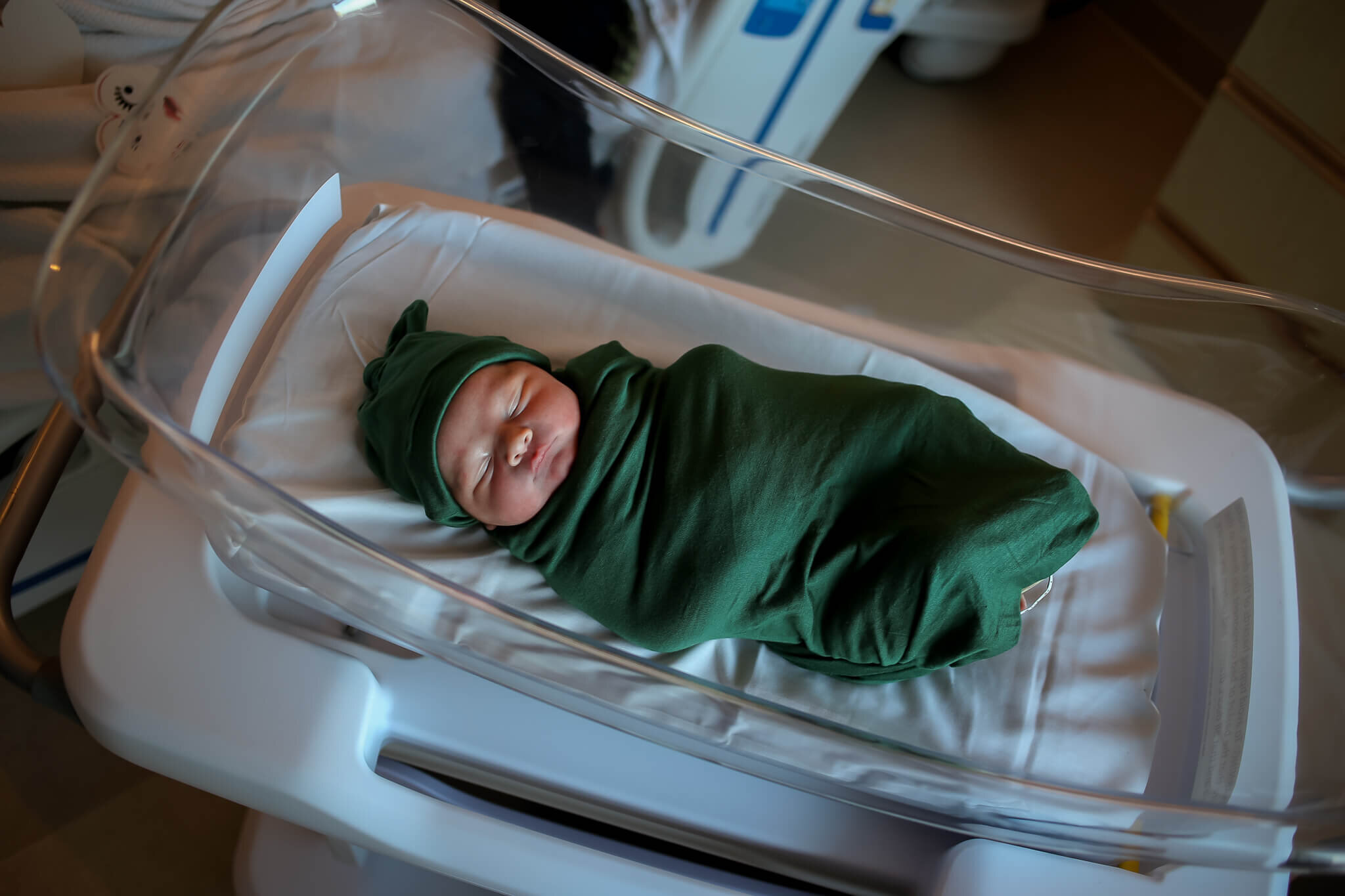 An image from above as the newborn baby lies in a hospital bassinet in his cap and tightly wrapped in a blanket from a session of fresh 48 baby pictures 