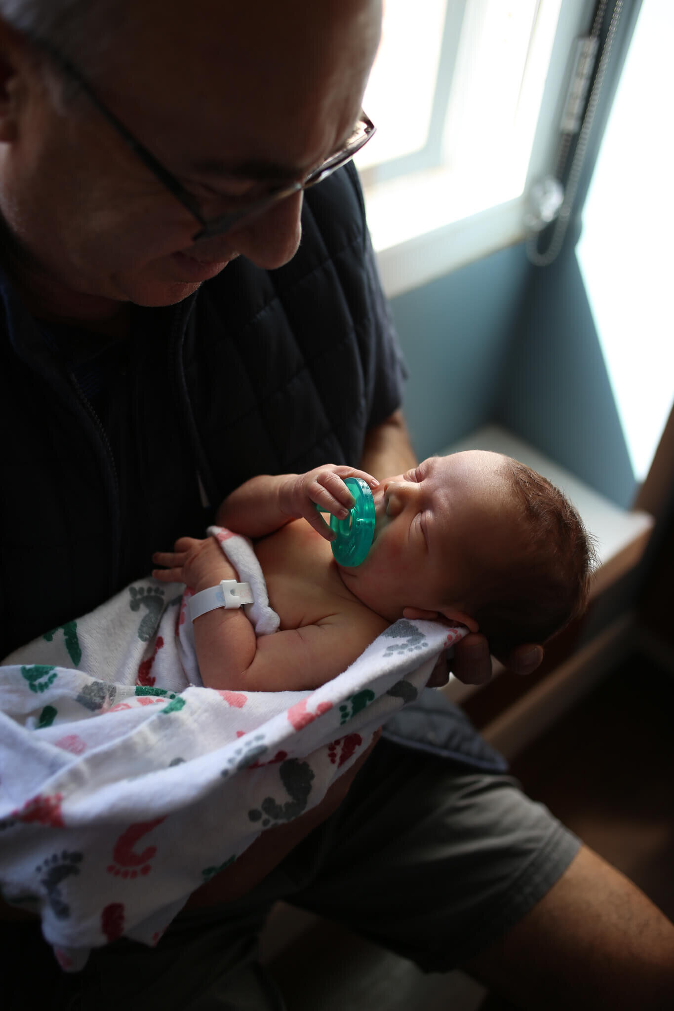  A picture of a new father holding his newborn child in his arms with the infant wrapped in a blanket and touching the pacifier in his mouth from a newborn session 