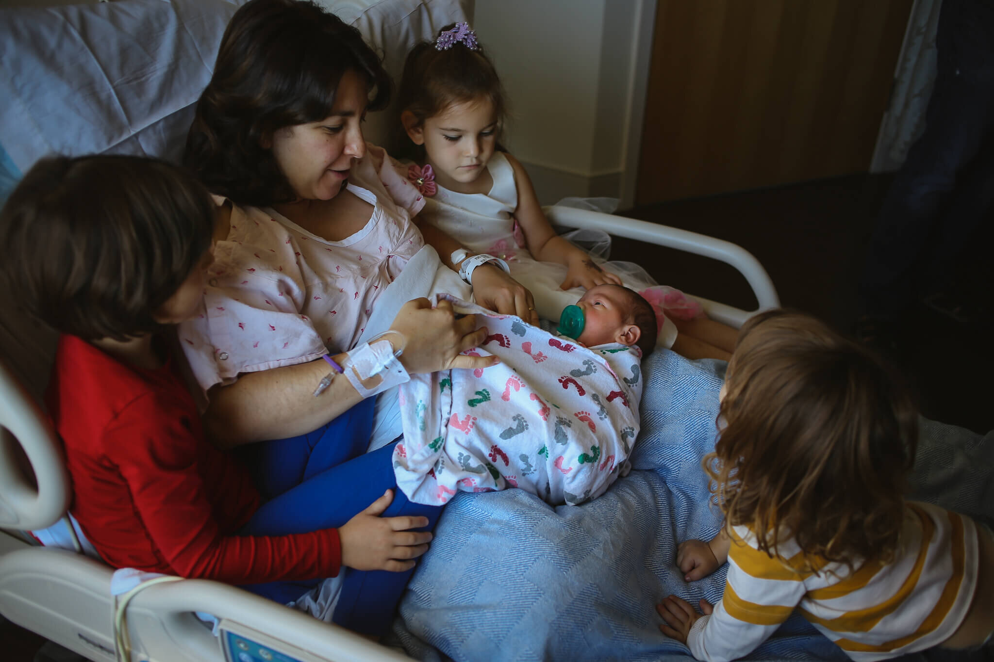  A photograph of mom in the recovery bed surrounded by her three children as they all gaze at the newborn on her lap, wrapped in a blanket and looking at mom with a pacifier in its mouth by Photography by L Rose - San Diego California newborn photogr