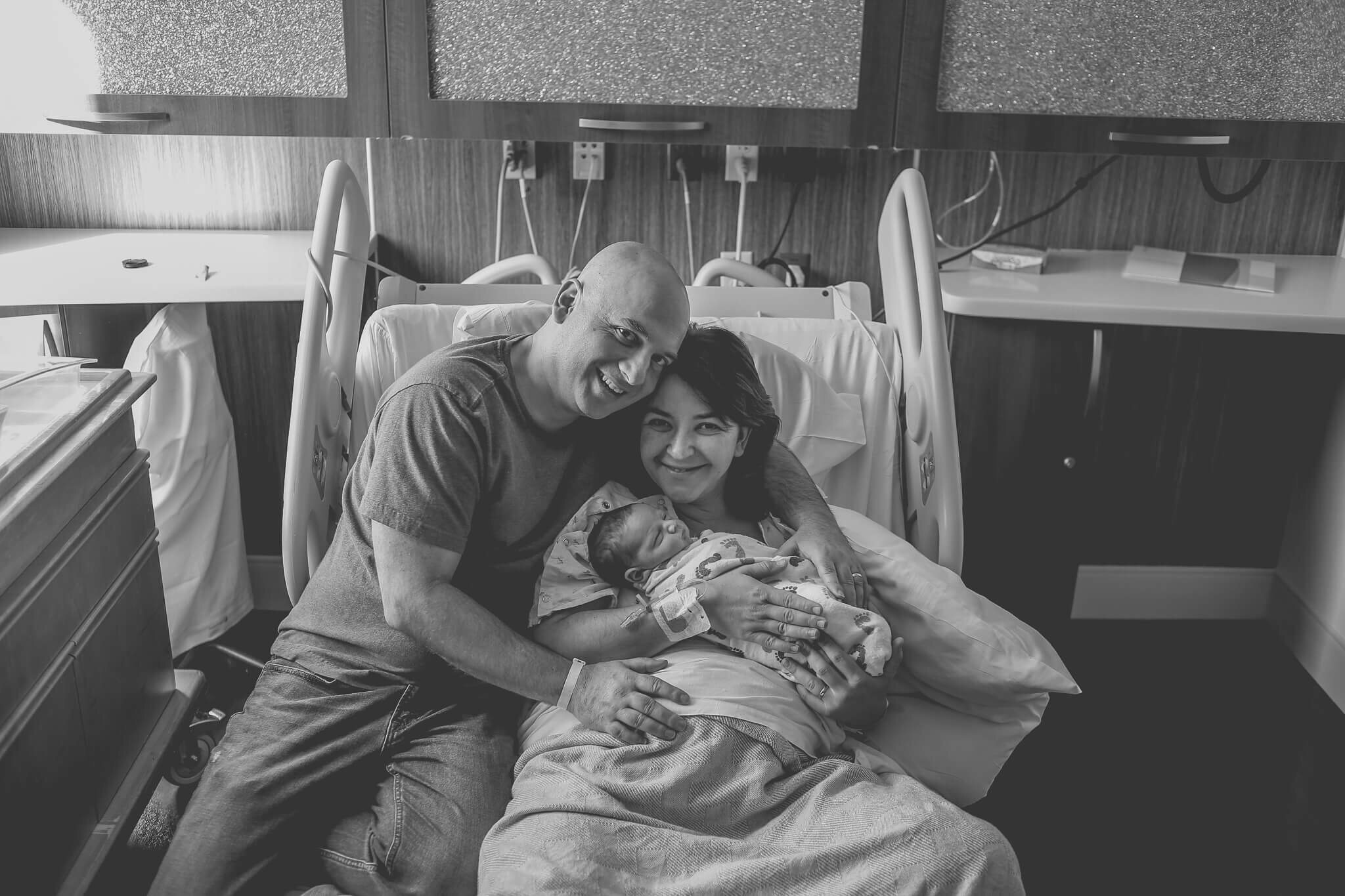  A photograph in black and white of a new father embracing his wife as she holds their newborn child while they all sit on the hospital recovery bed from a newborn picture session 