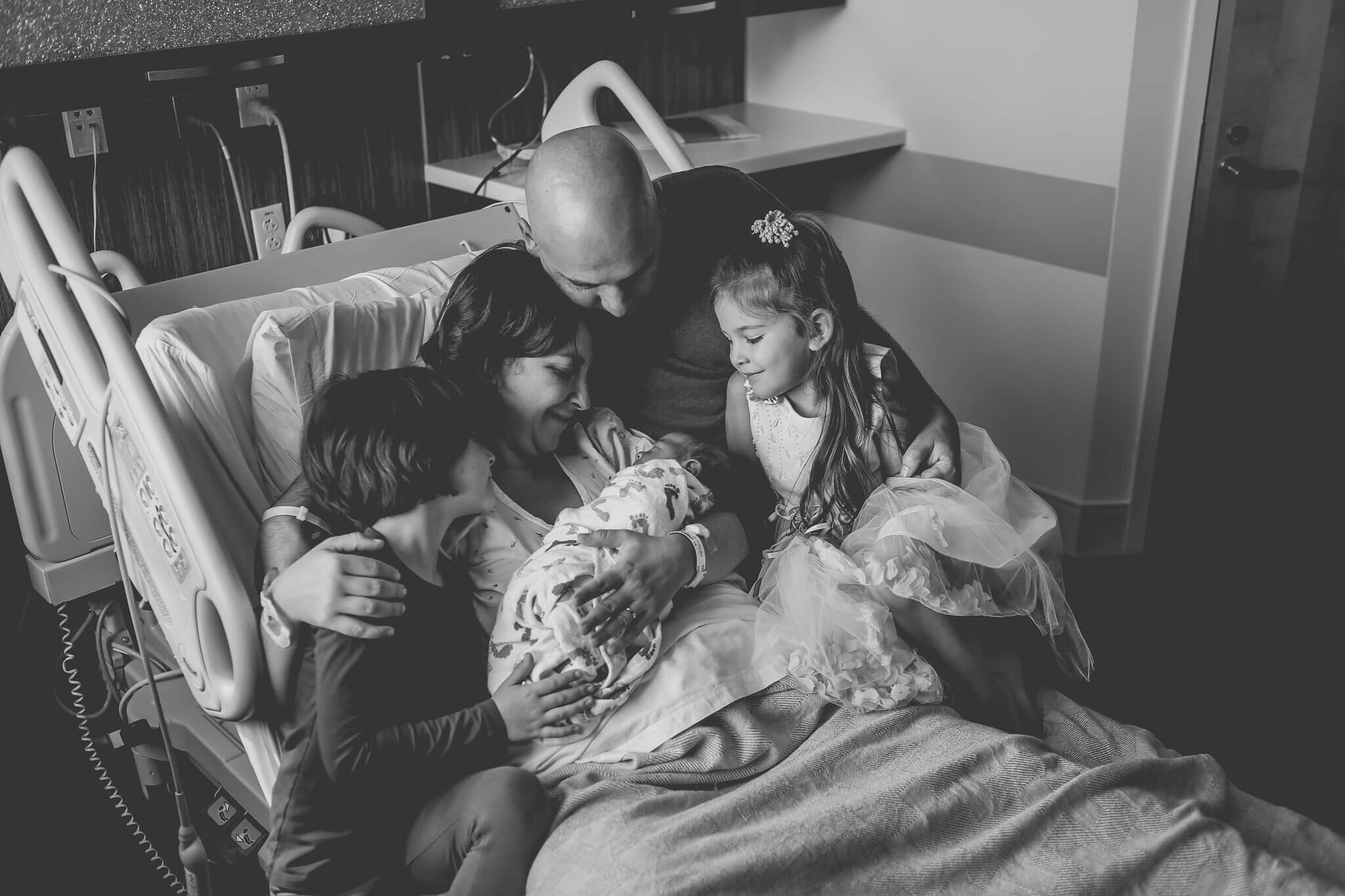  A picture in black and white of a family with dad and three older children gathered around the mother and their newborn baby who is wrapped in a blanket as they sit on the hospital recovery bed from a fresh 48 baby photo session 