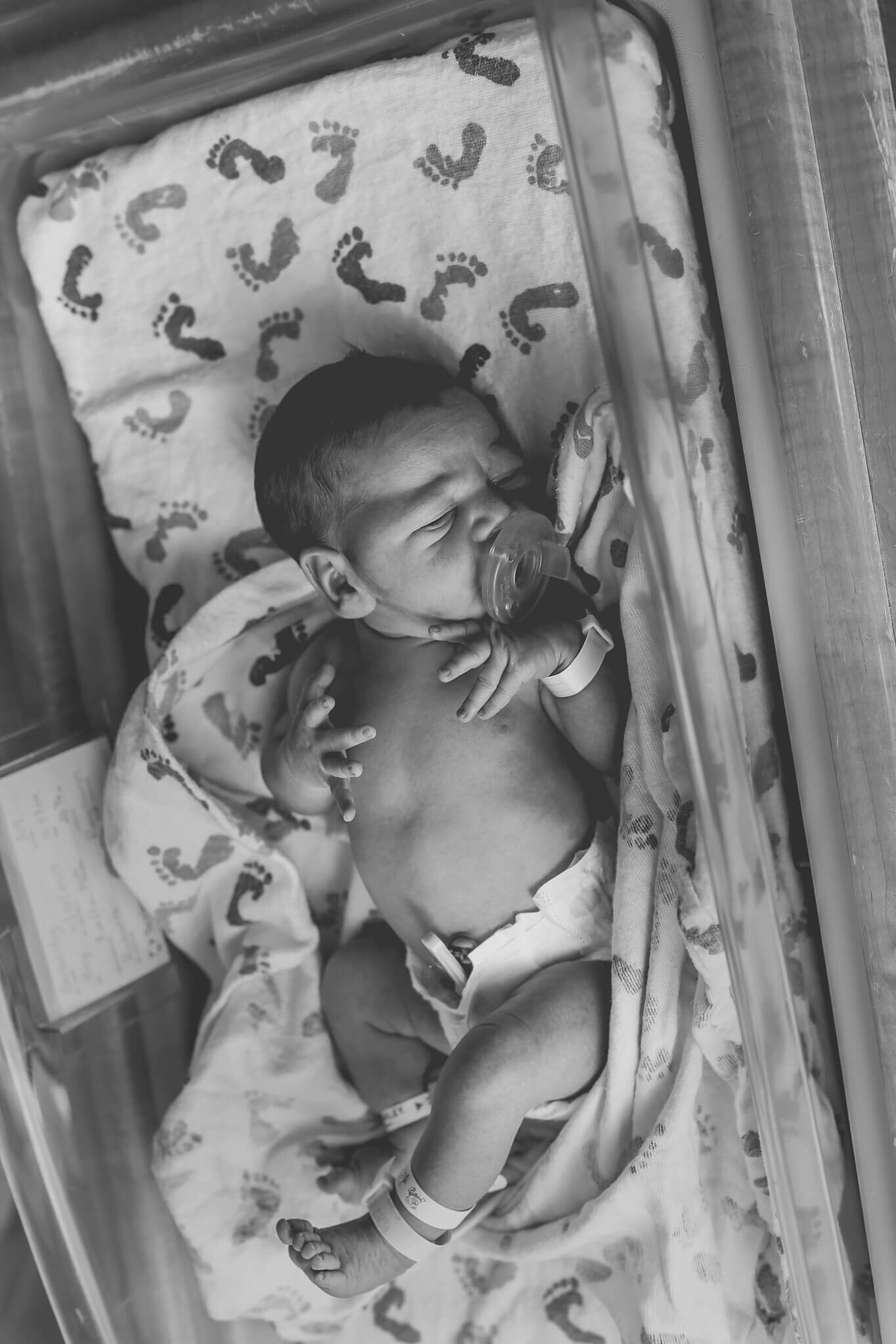  An image in black and white of a newborn baby lying in the hospital bassinet with an opened blanket, showing his tiny hands and belly and cord clamp by Photography by L Rose - San Diego newborn photography 