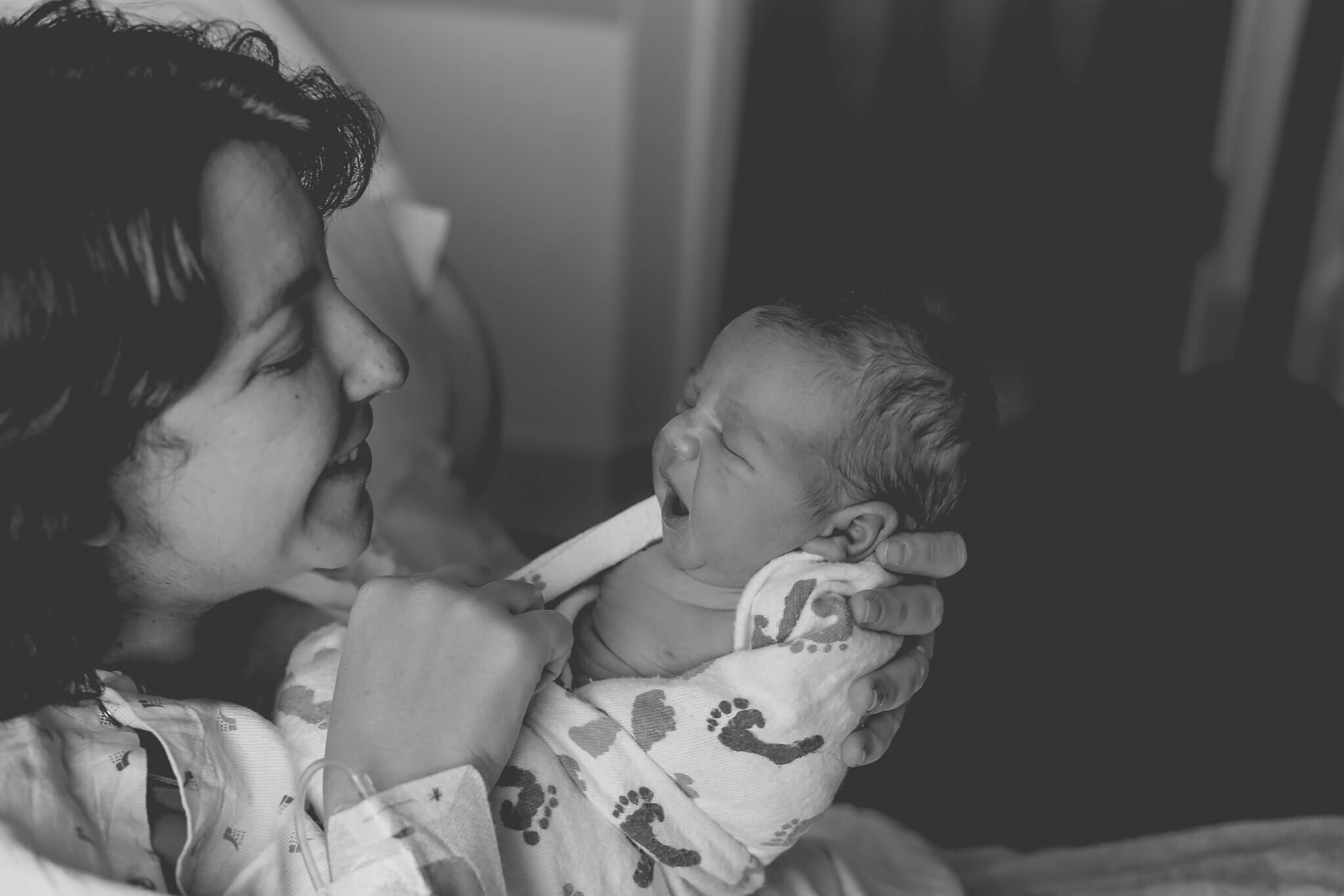  An image of a newborn yawning as mom holds him up close to her face while he is wrapped in a blanket by Photography by L Rose 