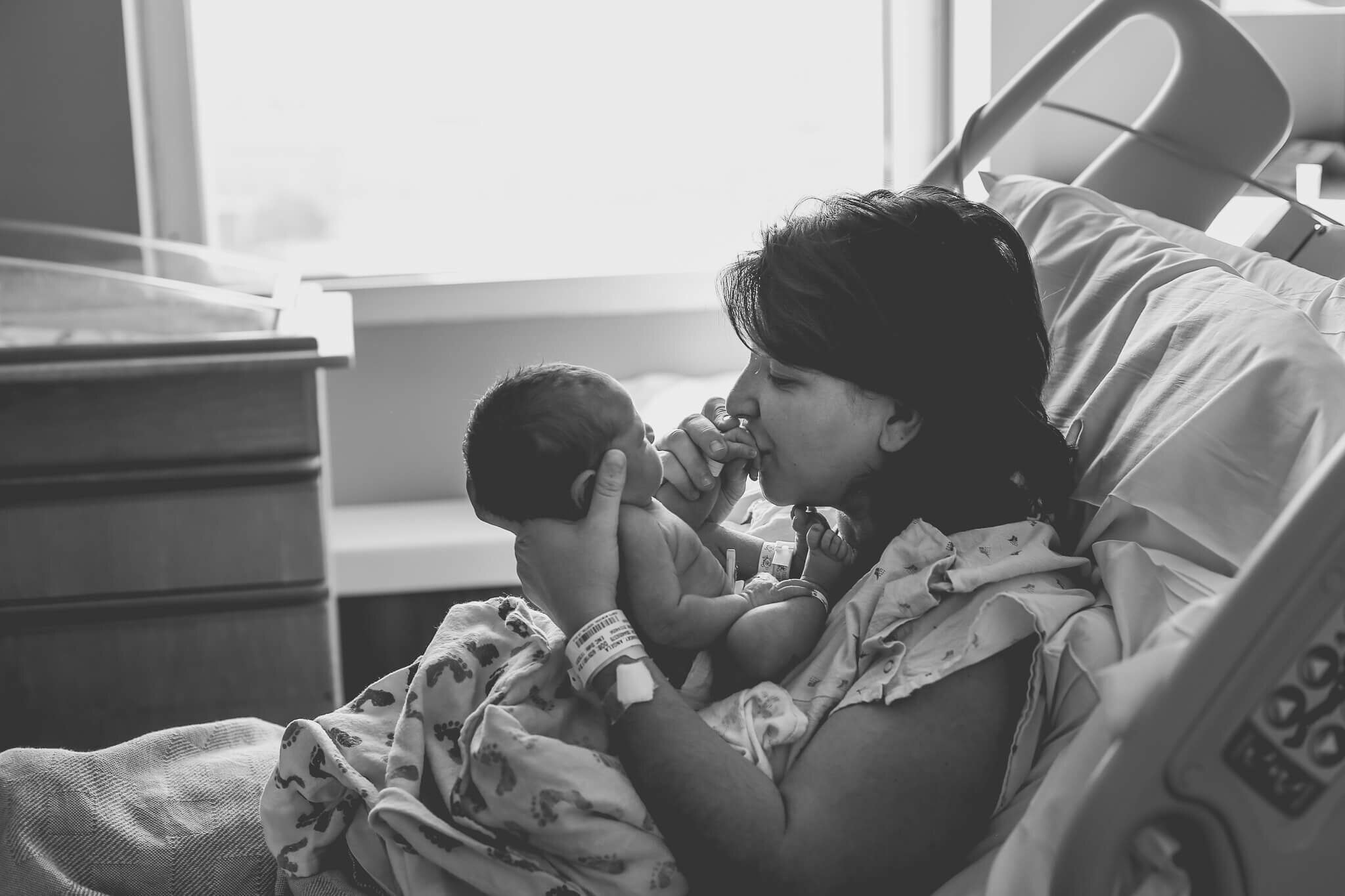  A photograph of a mom lying in her recovery bed, holding her newborn up in front of her, touching his bare skin and tenderly kissing his tiny hand by Photography by L Rose 