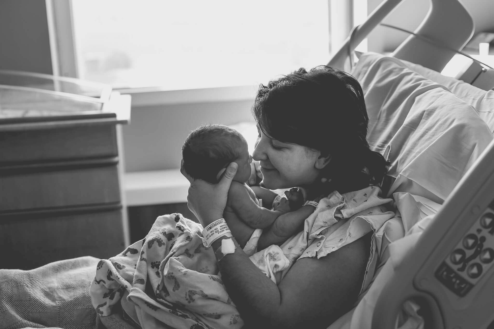  A picture of a new mother sitting in her delivery bed, holding her newborn up close to her face as she leans forward to touch noses with him by Photography by L Rose 