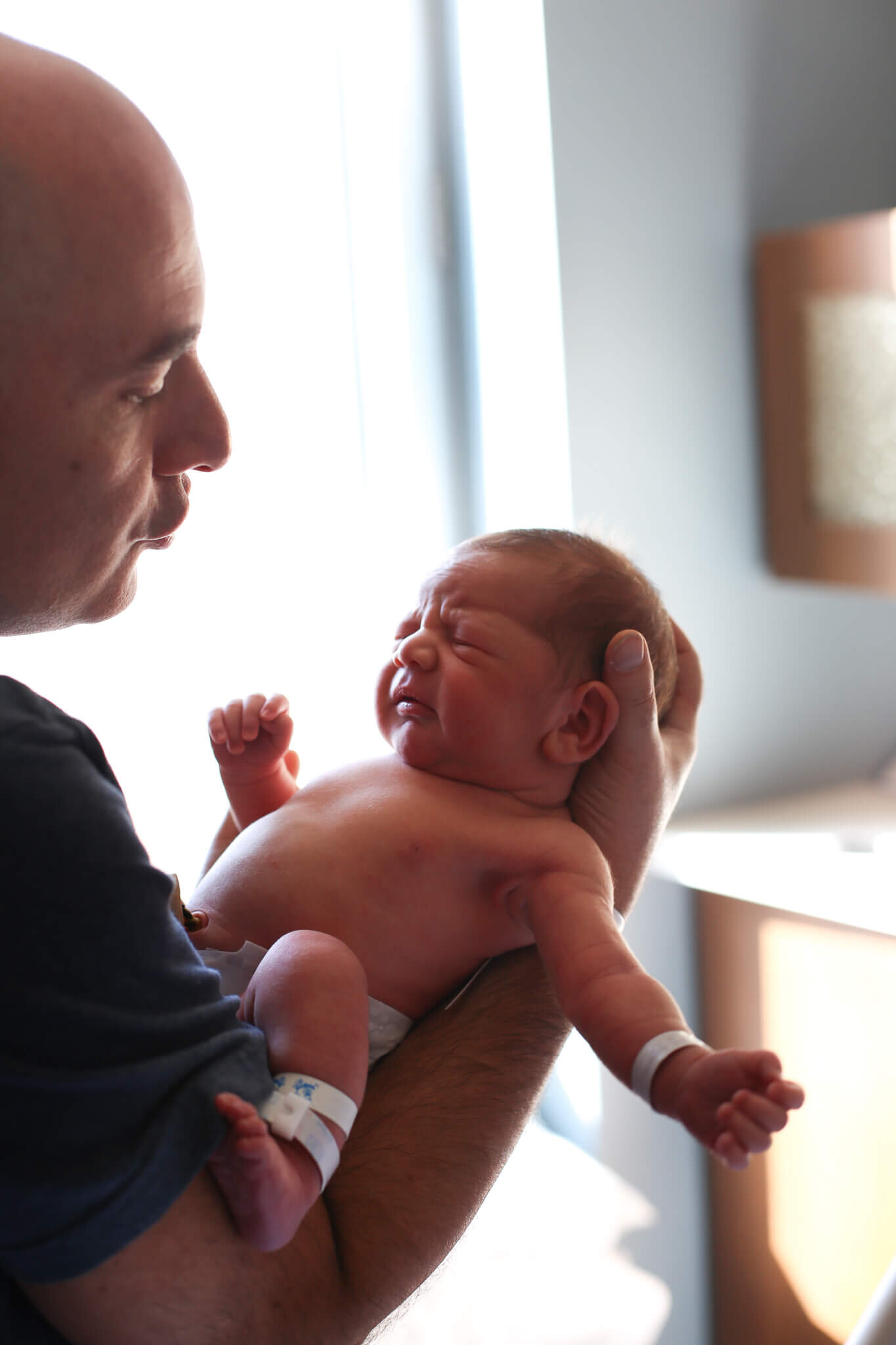  An image of a new dad holding his newborn in his hands, trying to quiet the baby as the baby starts to cry by Photography by L Rose 