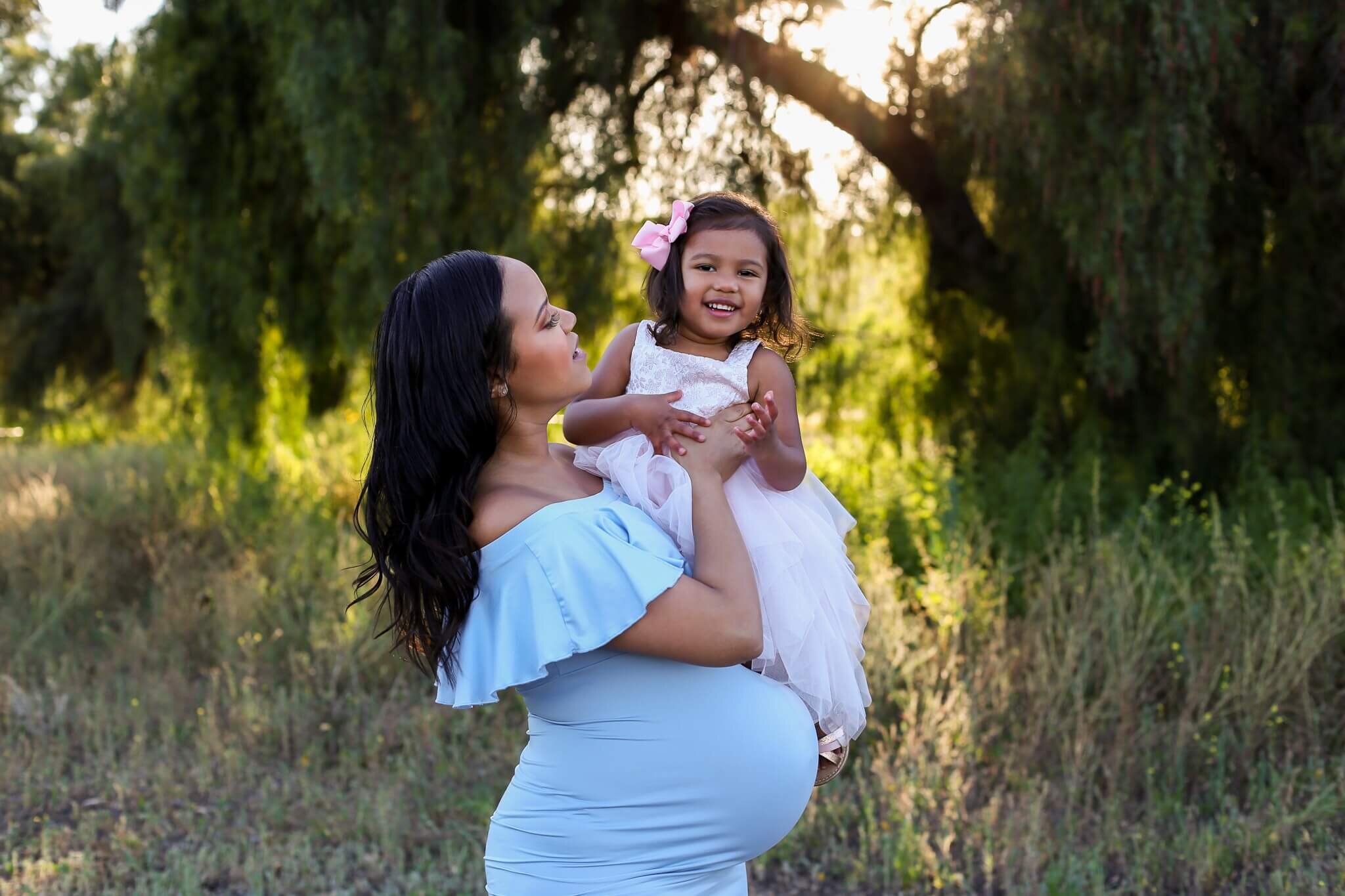  A picture of a pregnant mom holding her young daughter who smiles and enjoys the family time together in a park with sunlight and trees behind them by Photography by L Rose 