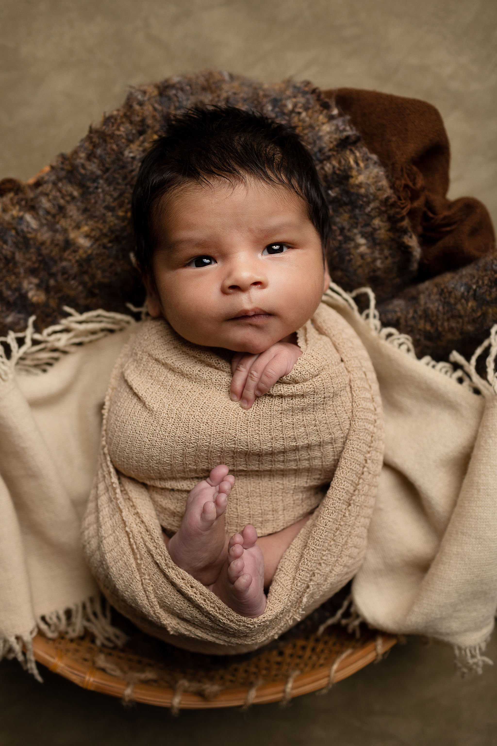  baby with wide-open eyes looking into camera wth little feet and toe poking out of the blanket 