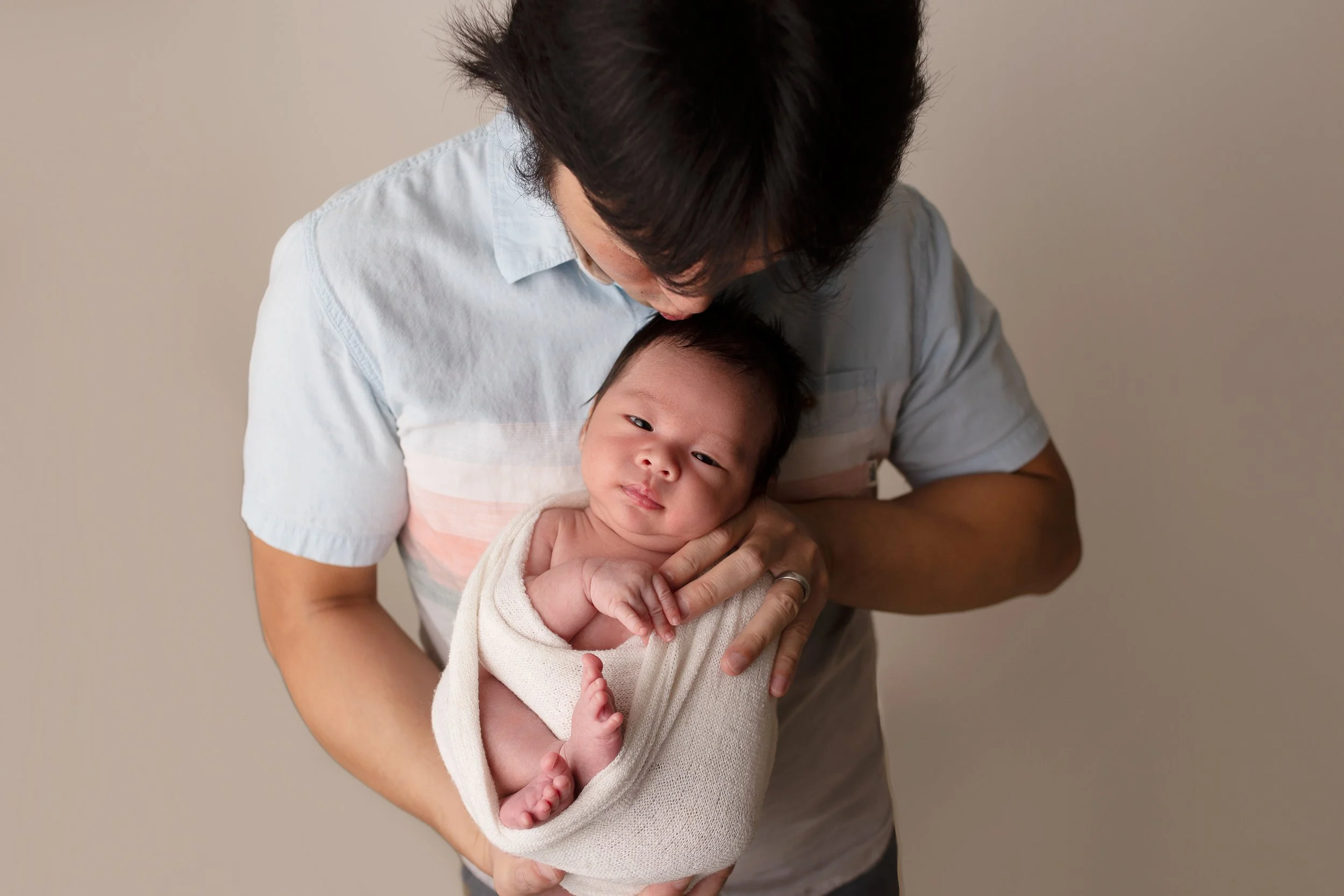 newborn photo session with the father holding the baby and kissing his hed while baby is wrapped in a white blanket