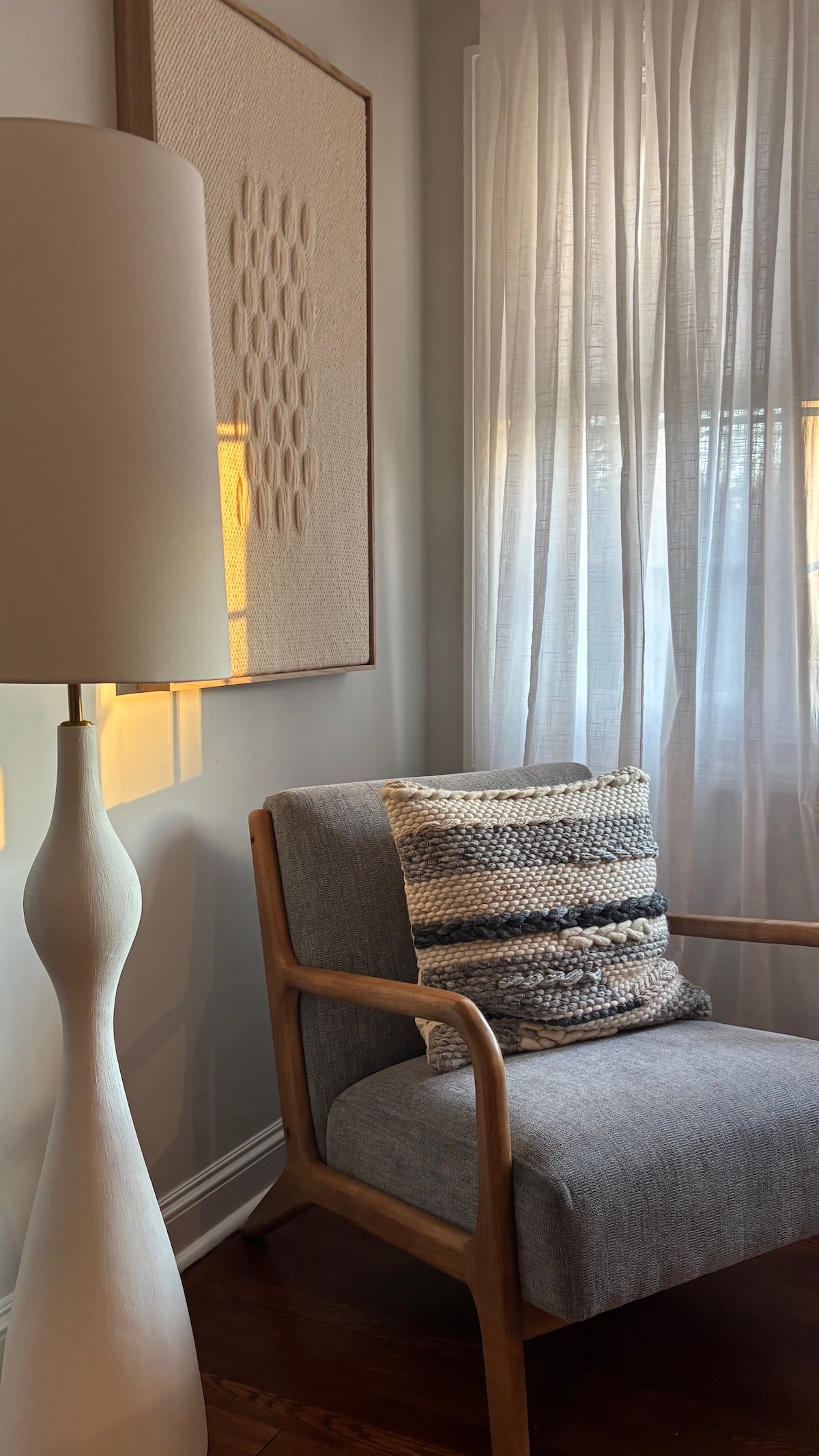 Wood-framed accent chair with a woven pillow beside a white floor lamp, textured wall art, and sheer curtains, design consulting in Westchester, NY