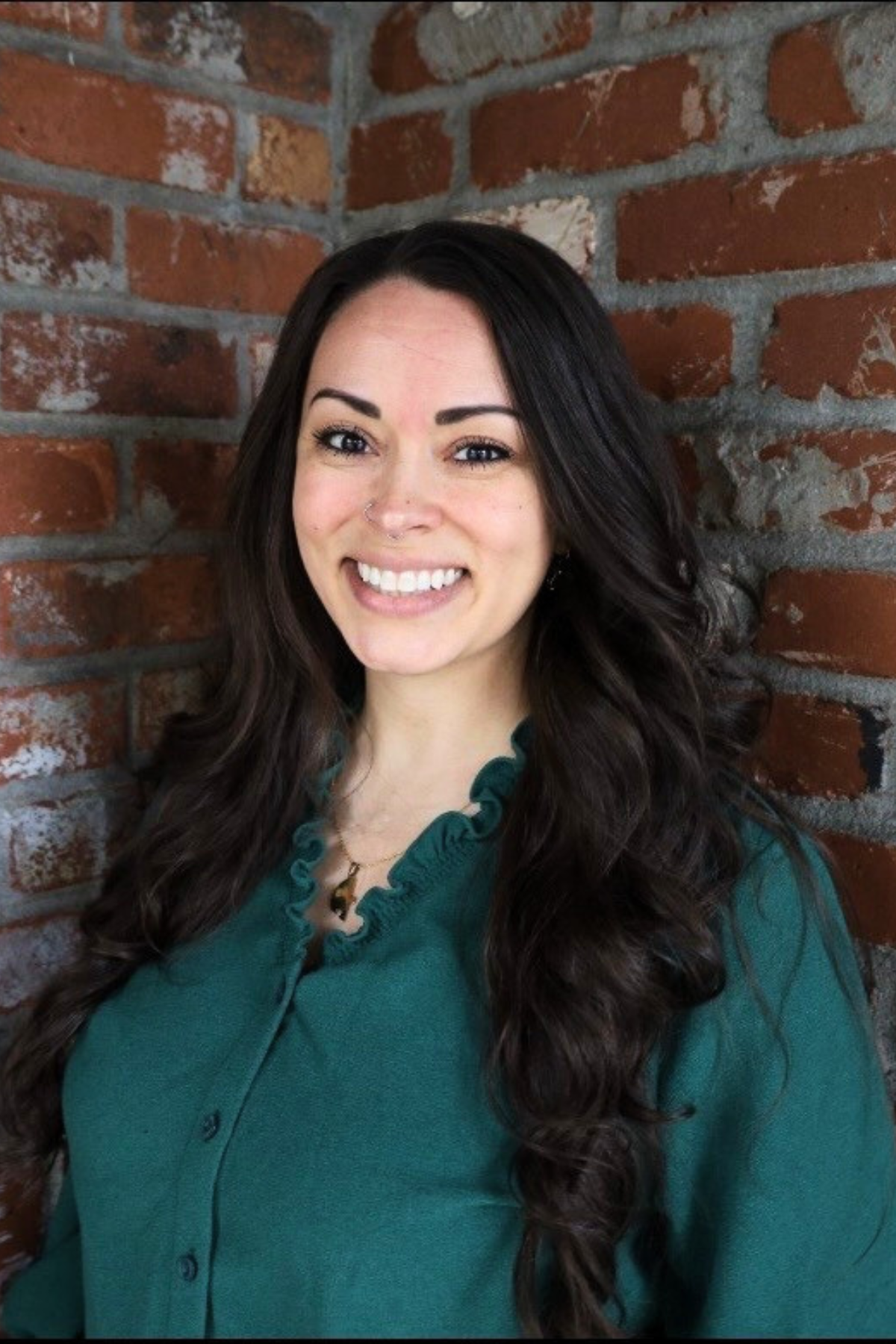 A headshot of Krystle May who has long dark hair and smiles in front of a brick wall.