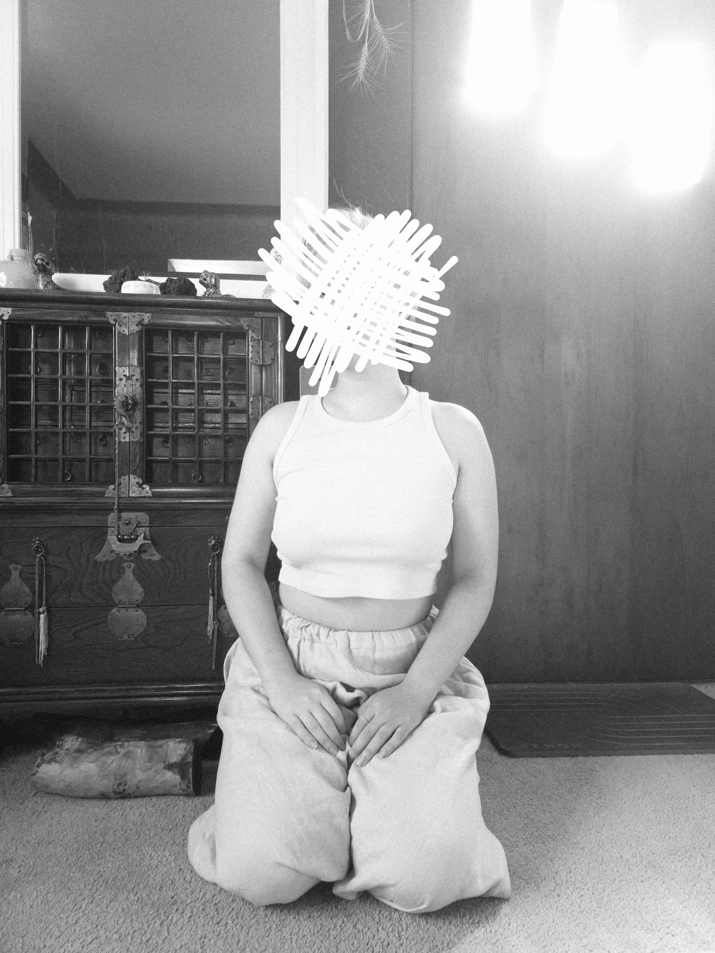 A black and white photo of a person kneeling on a floor. Their face is scratched out with crosshatched white lines.