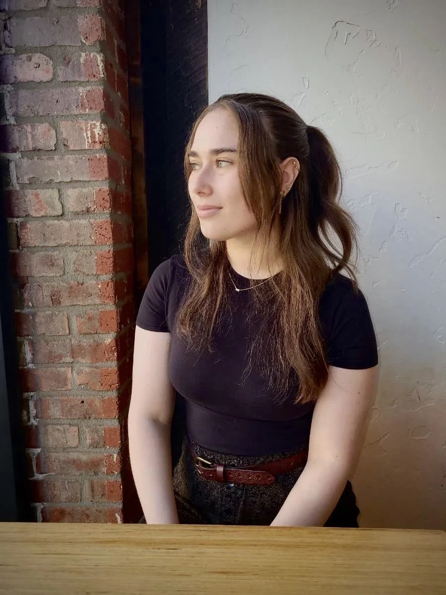 This is a photo of artist Kayla Reese. She stands in front of a brick wall. She has long, brown hair and is wearing a black t-shirt.