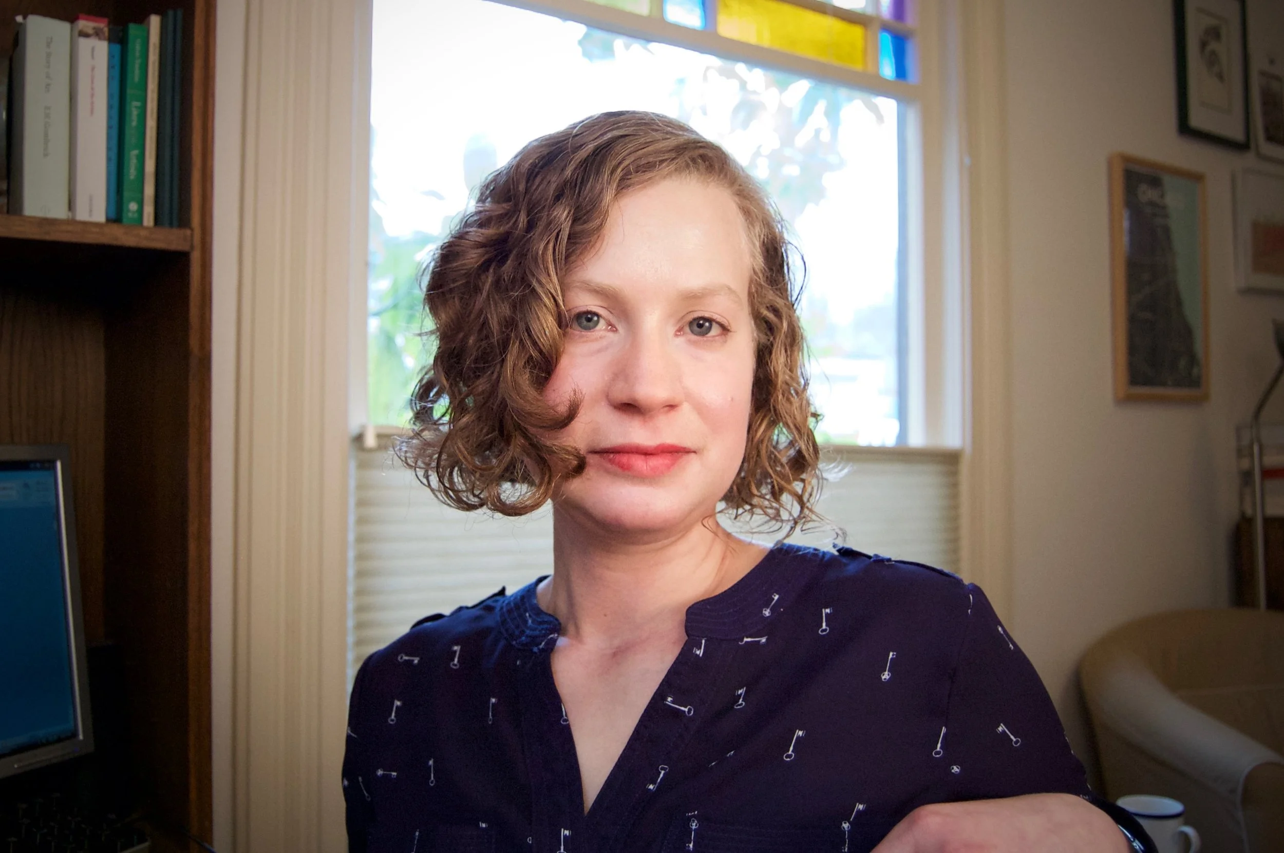 This is a photo of author Anca L. Szilágyi. She has curly blonde hair and is standing in the sunlight from a window in her home.