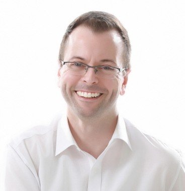 This is a photo of author Billie Pritchett. He has dark blonde hair, glasses, and is wearing a white, collared shirt. He is smiling.