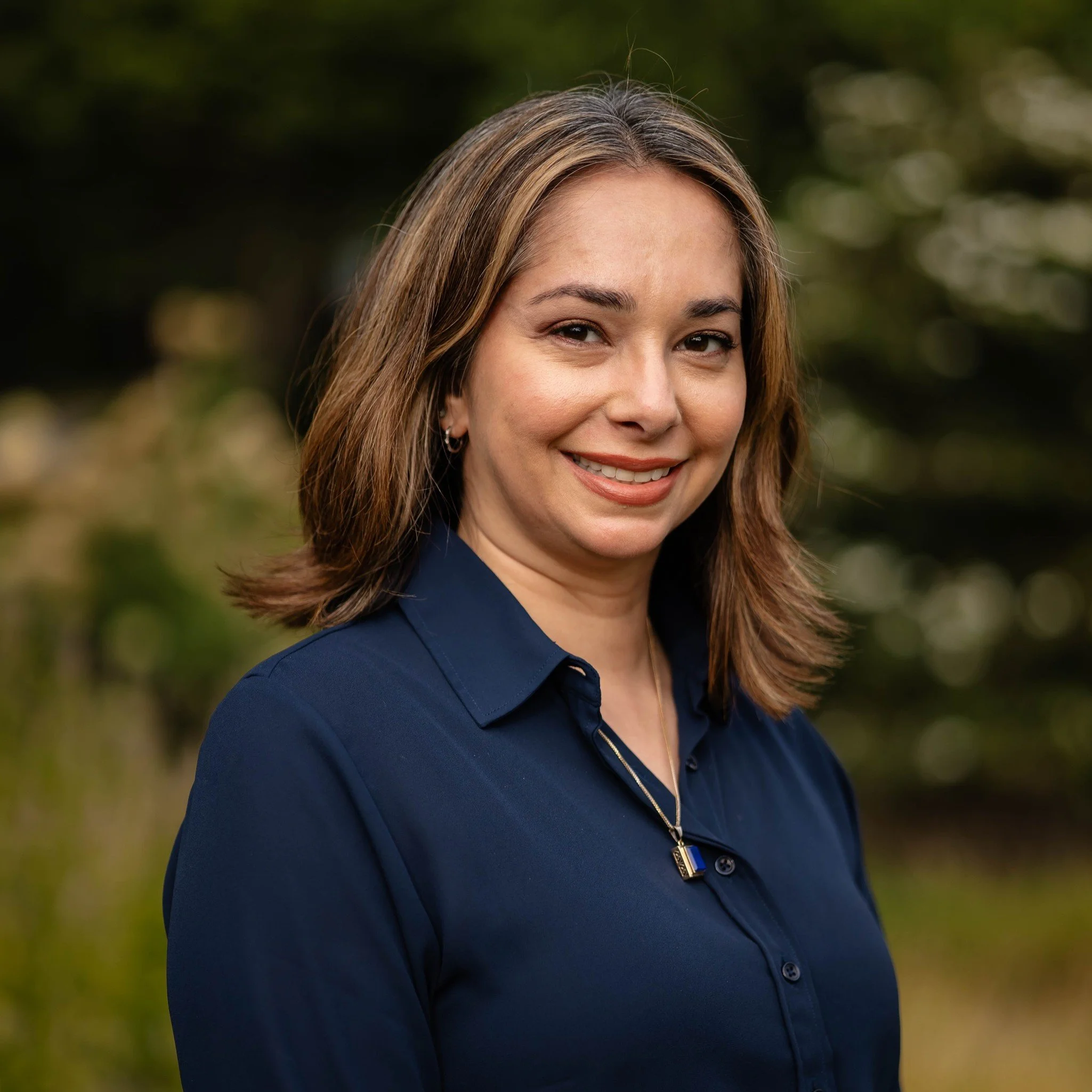 Professional woman with long dark hair smiling, wearing a white collared shirt and a black blazer, against a neutral background.
