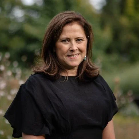 A woman with shoulder-length dark hair, wearing a dark blazer and a blue top, smiling at the camera against a dark background.